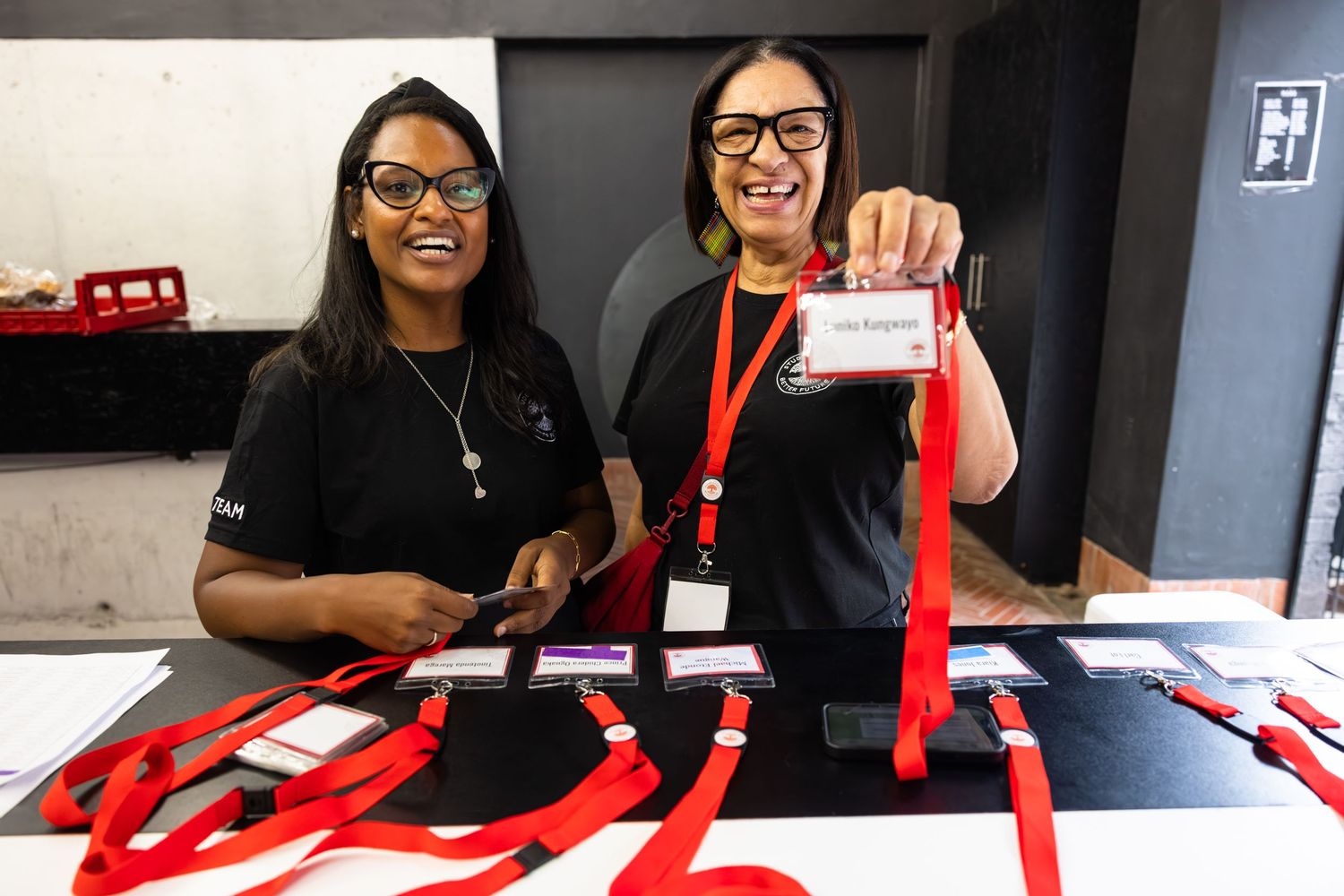 Two staff members stand at a registration desk with red lanyards and event materials.
