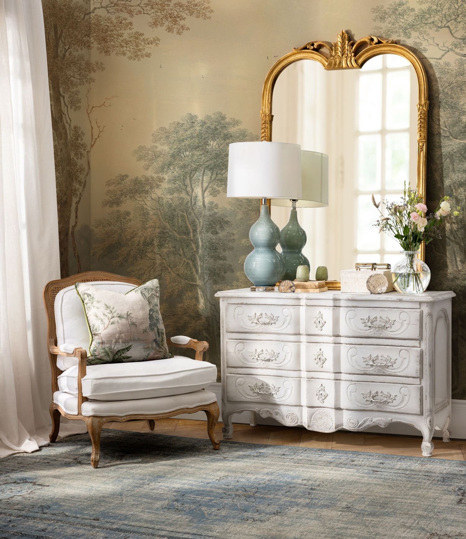 Elegant French country living room with ornate gold mirror, white dresser, blue accents and scenic wallpaper mural.