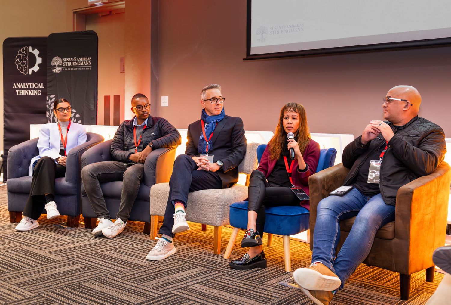 A row of professionals seated on orange chairs during a conference presentation.