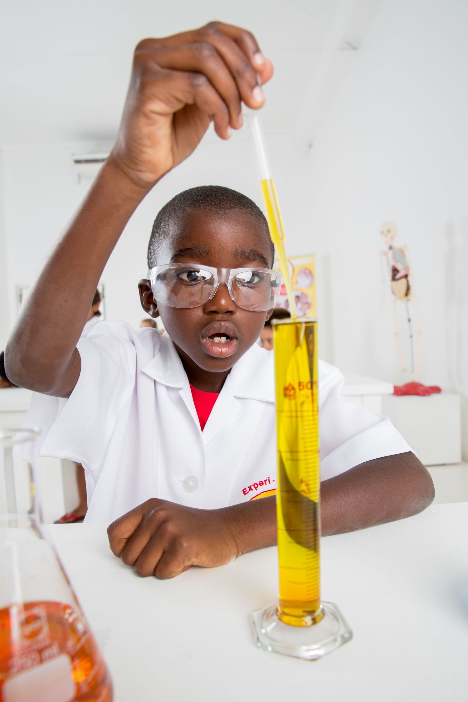 Student wearing protective goggles measures yellow liquid in graduated cylinder during lab experiment.
