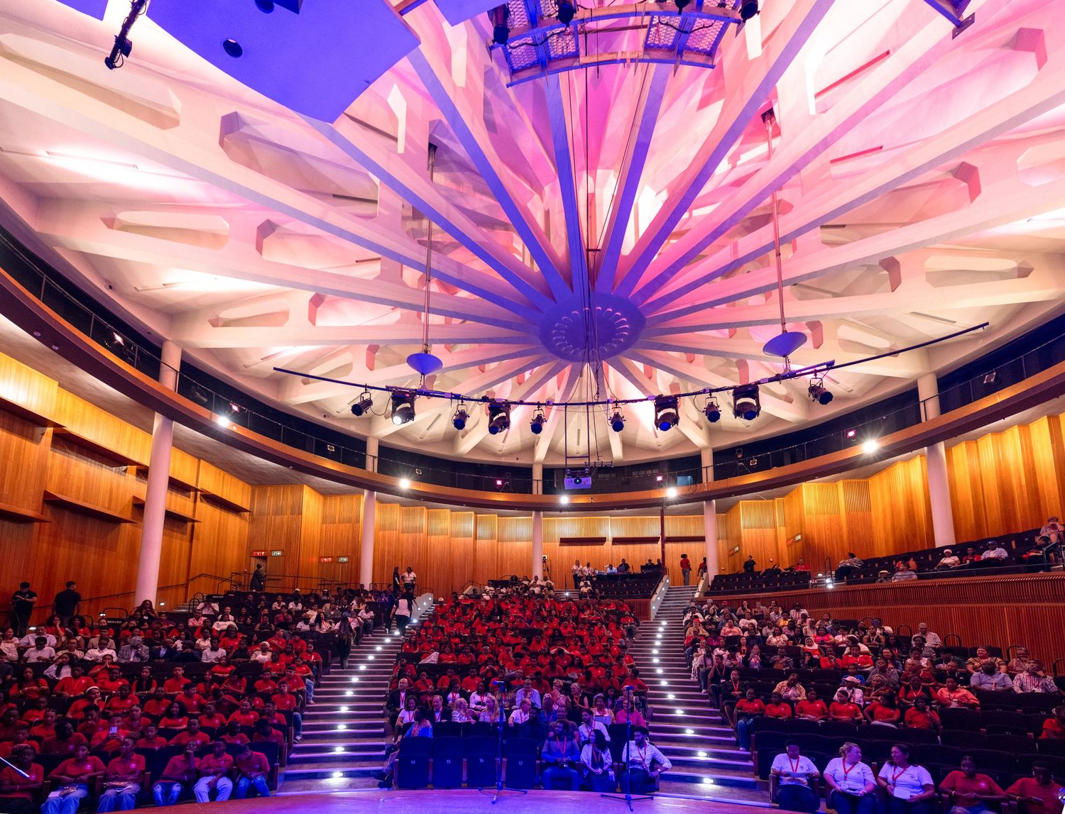 Large auditorium with dramatic purple and pink lighting illuminating a domed ceiling during an event.