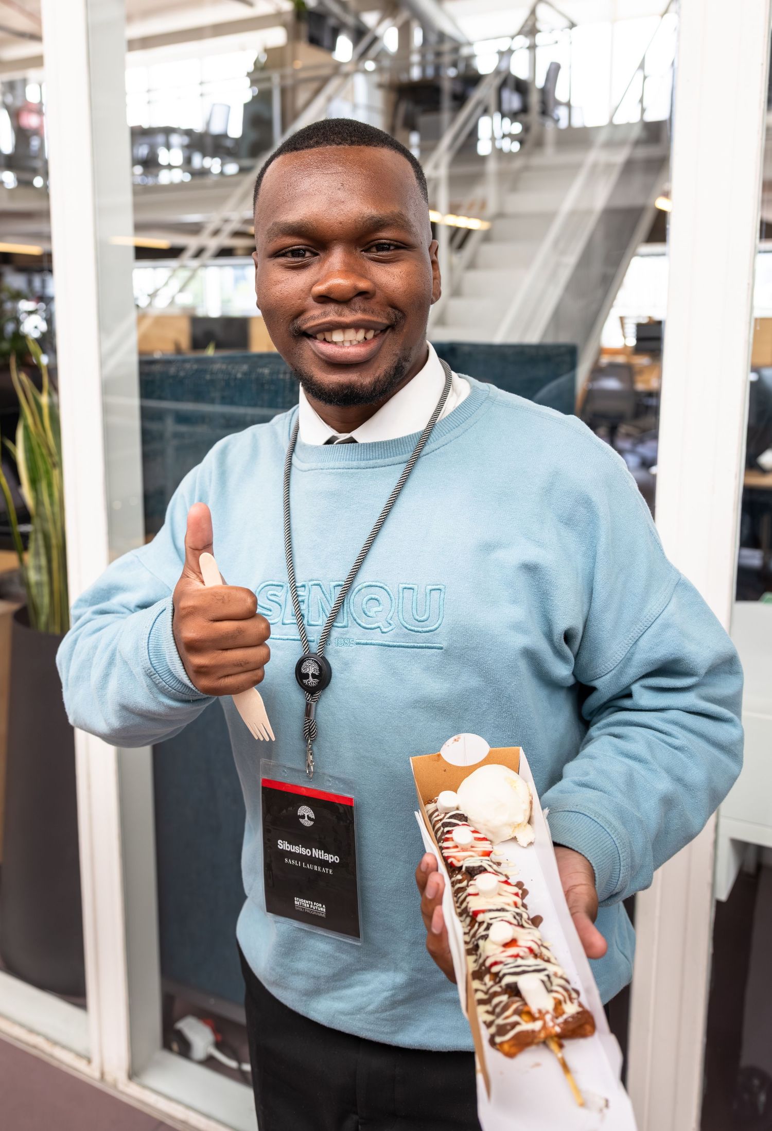 Event attendee in light blue sweater gives thumbs up while holding a snack bar at indoor venue.