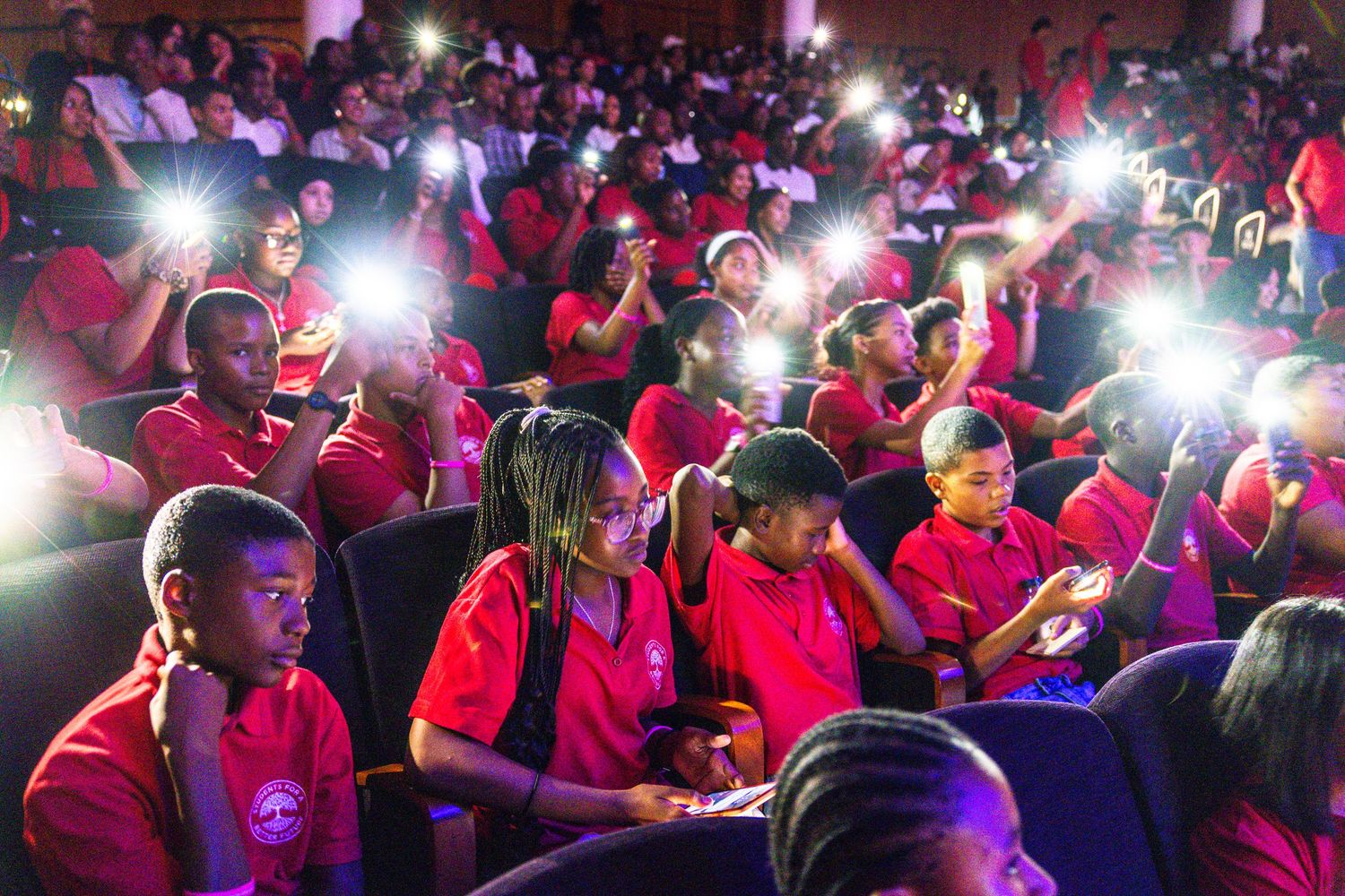 A crowded event venue with red-shirted attendees and camera flashes illuminating the dark space.