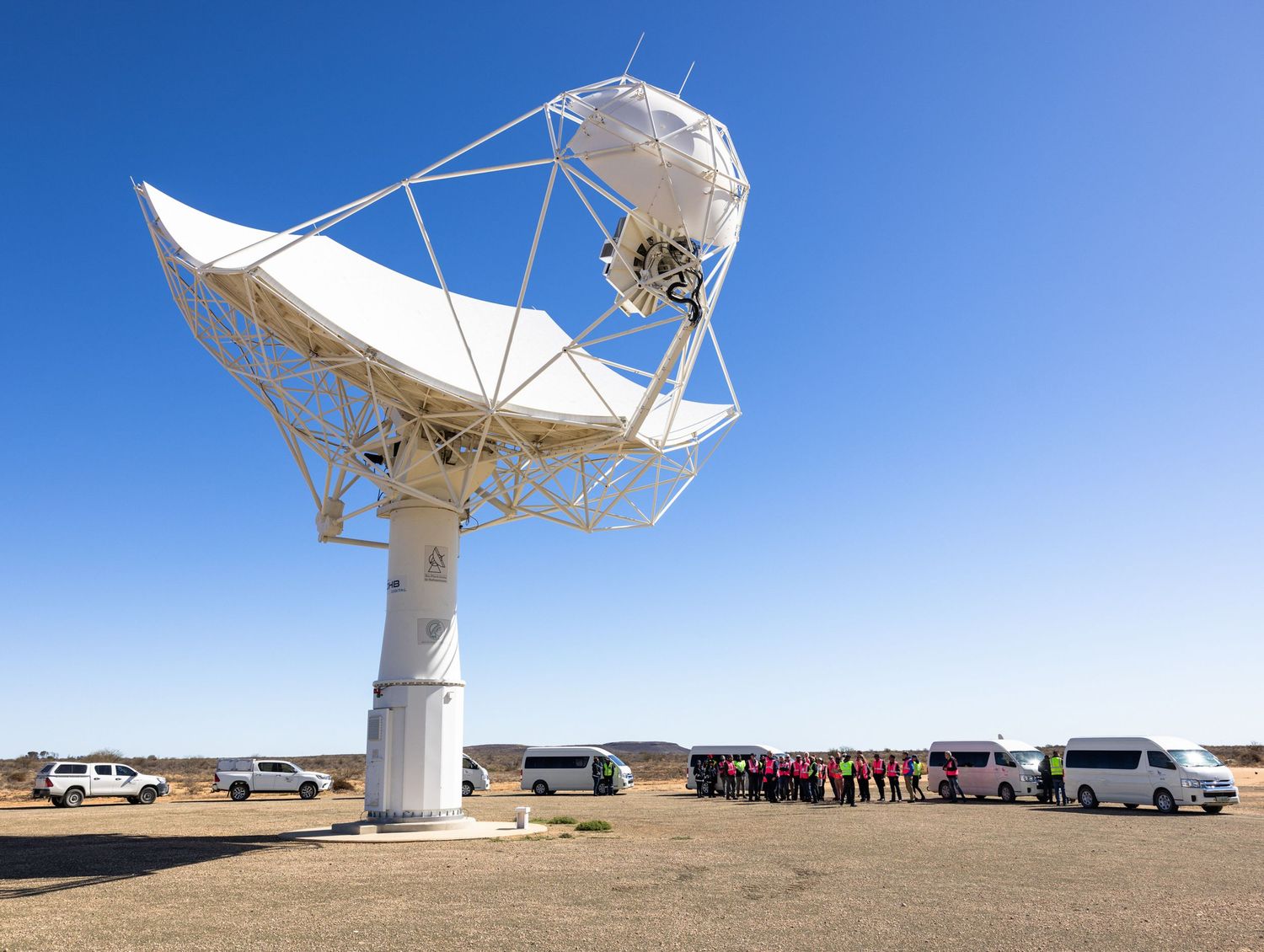 White radio telescope dish installation with parked vehicles and tour groups visible in desert setting.