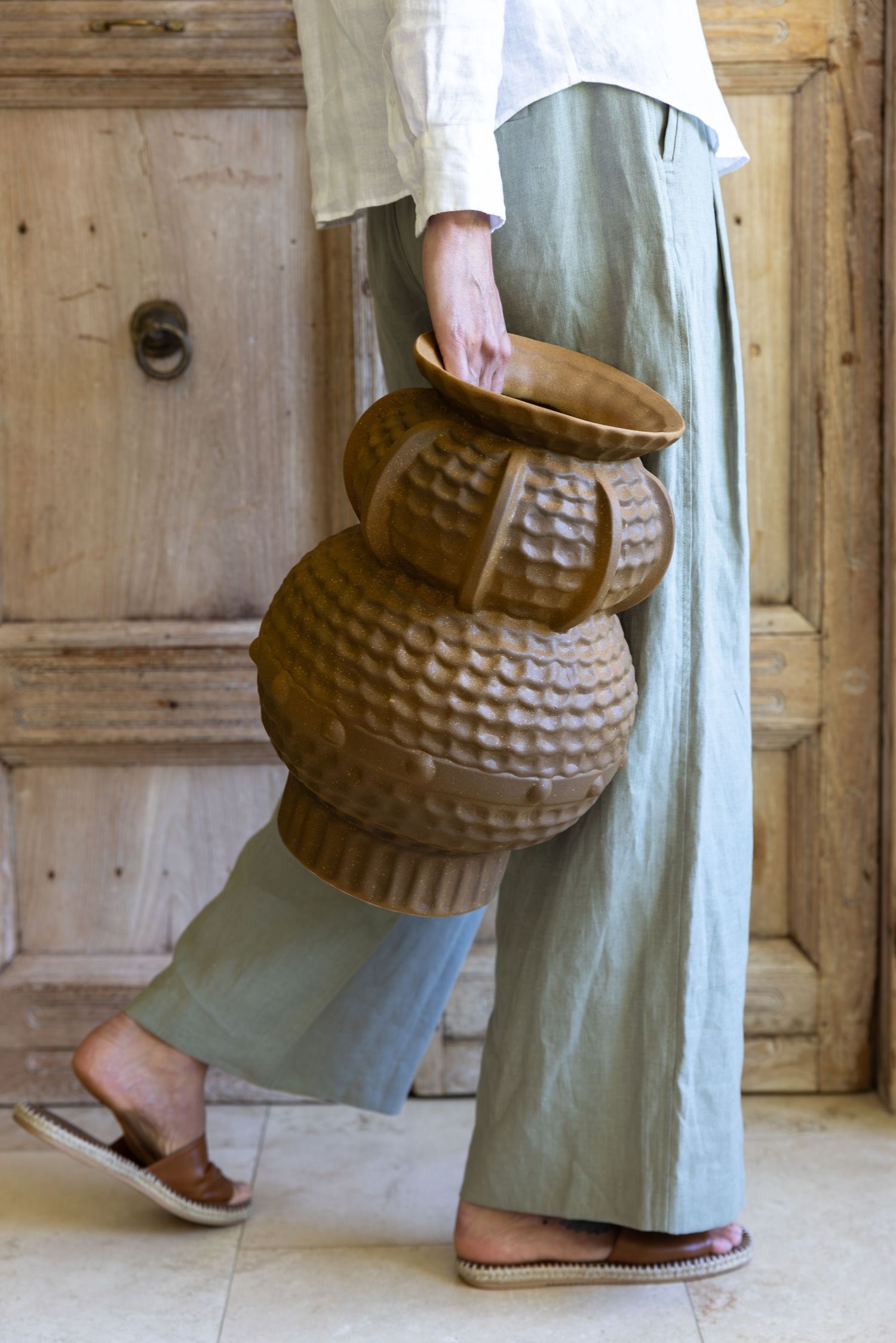 Person in linen clothing carrying large textured ceramic vases against a rustic wooden door.