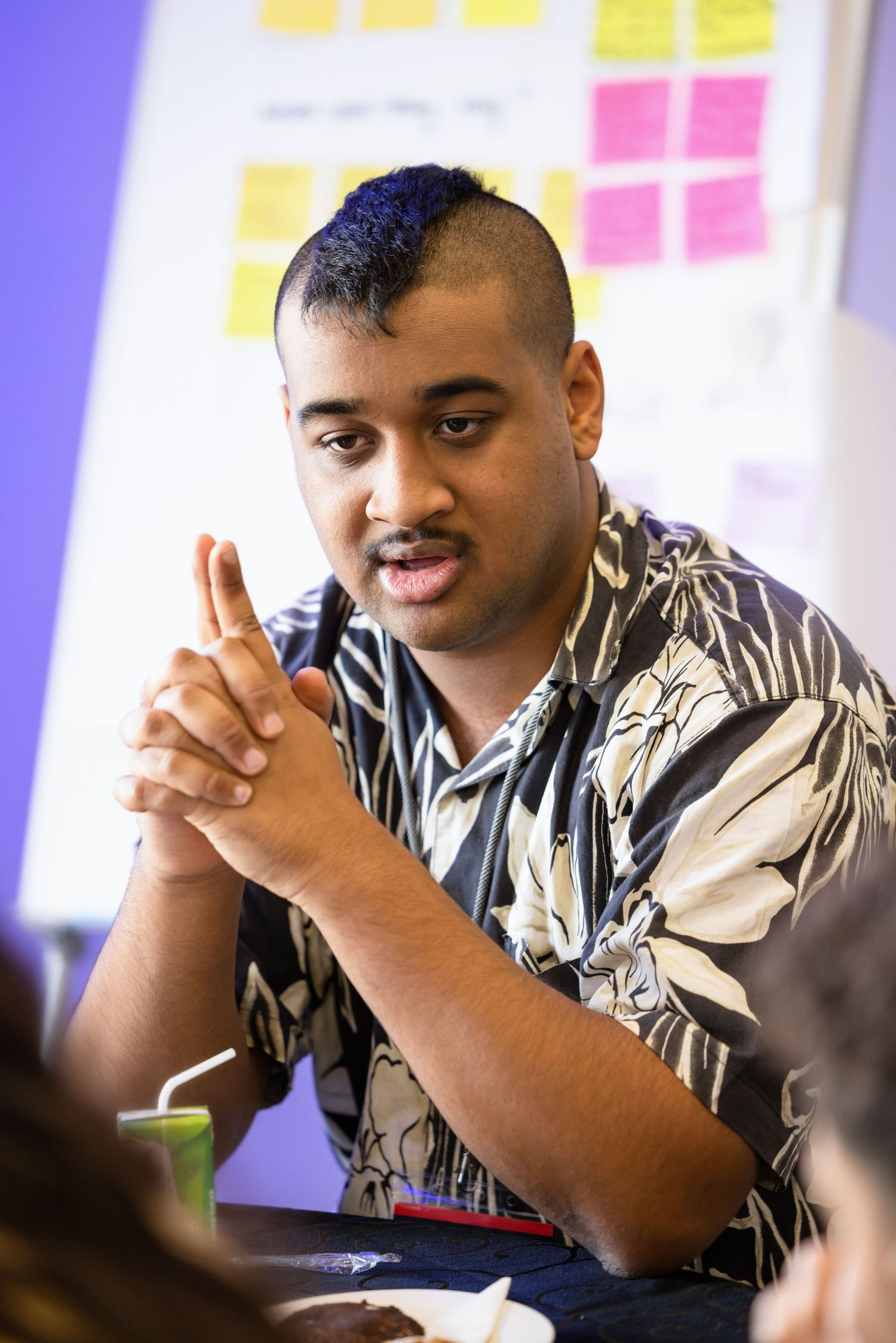 Professional in floral shirt speaks during a meeting with sticky notes on wall in background.