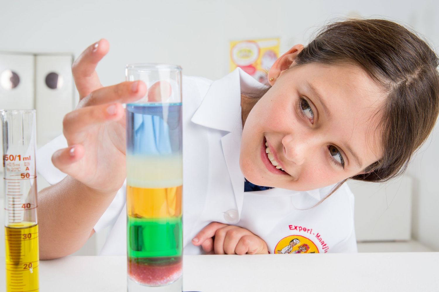 Student in white lab coat observes layered liquids in test tube during science class.