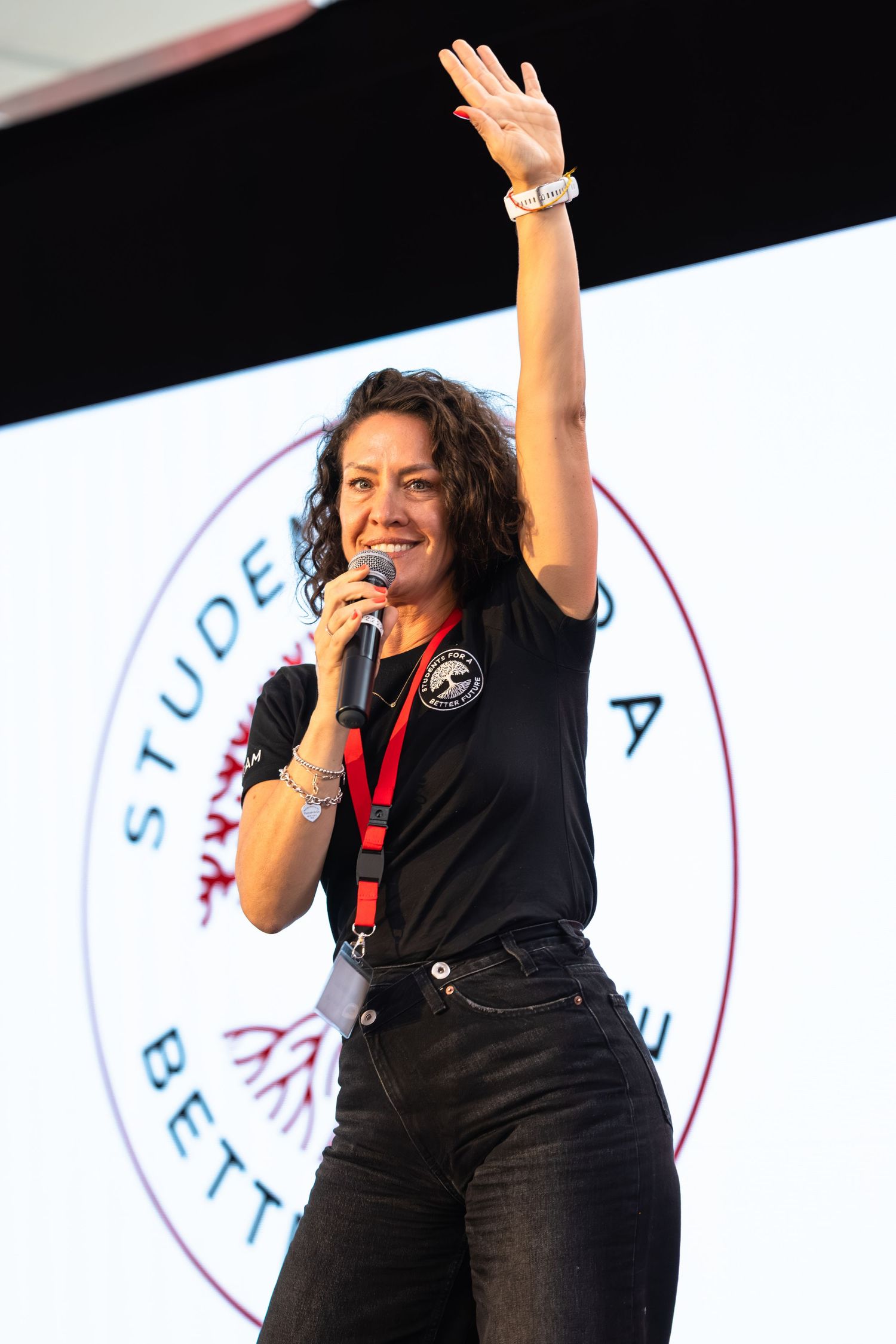Speaker in black outfit raises arm while speaking into microphone at event with circular logo backdrop.
