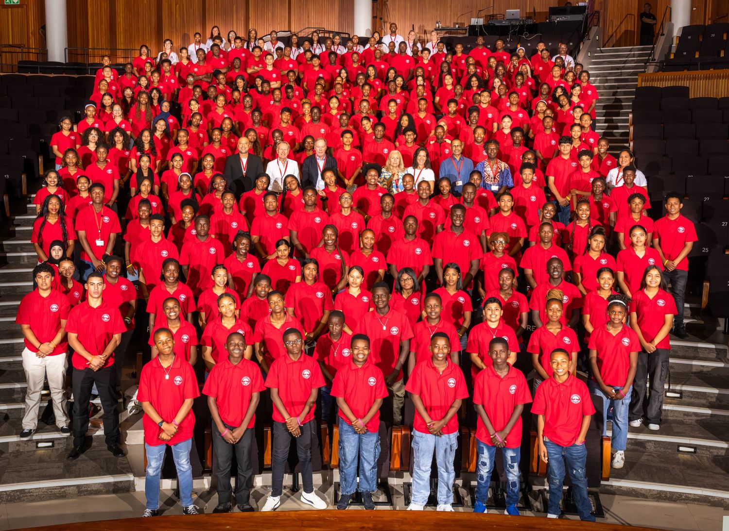 Large group photo of people wearing matching red shirts posed in rows on auditorium steps.