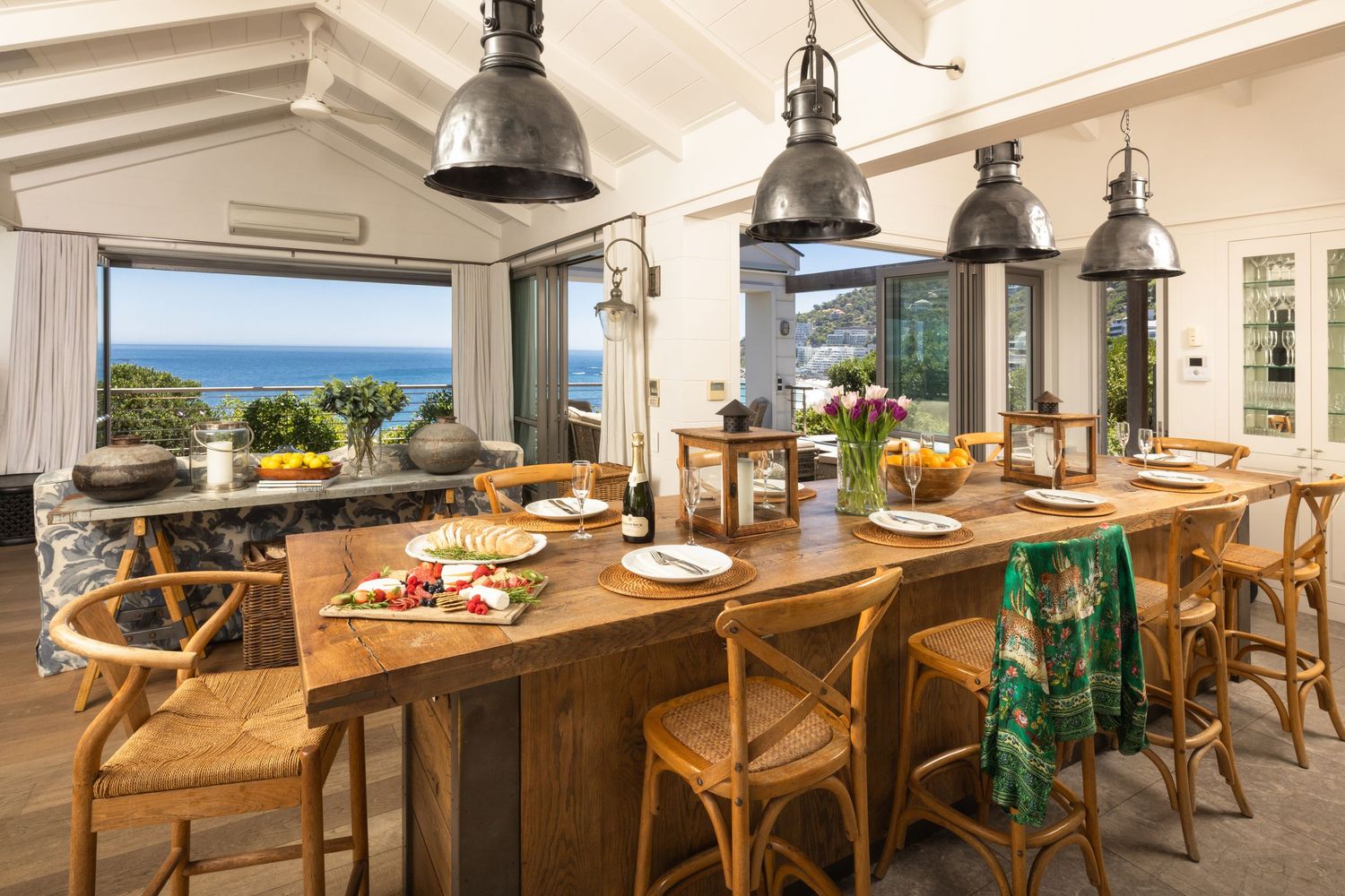 Rustic dining area with wooden table and chairs under industrial pendant lights with ocean views.