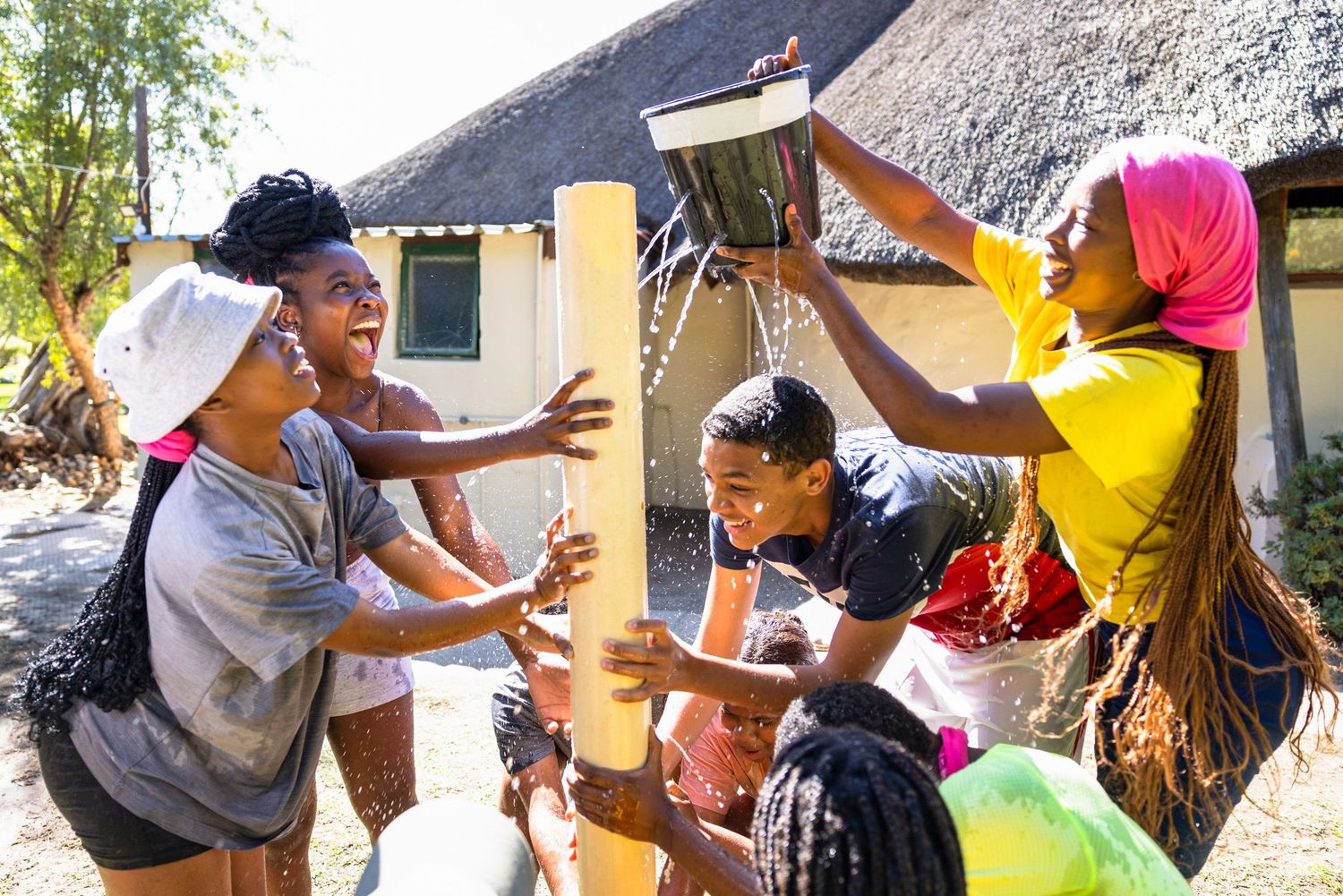 Group plays energetic outdoor game with colorful equipment near thatched building.