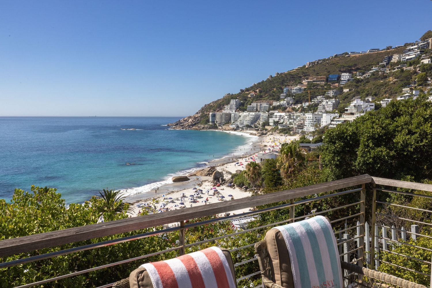 Coastal view from a balcony showing white buildings cascading down a hillside to a pristine beach and blue ocean.