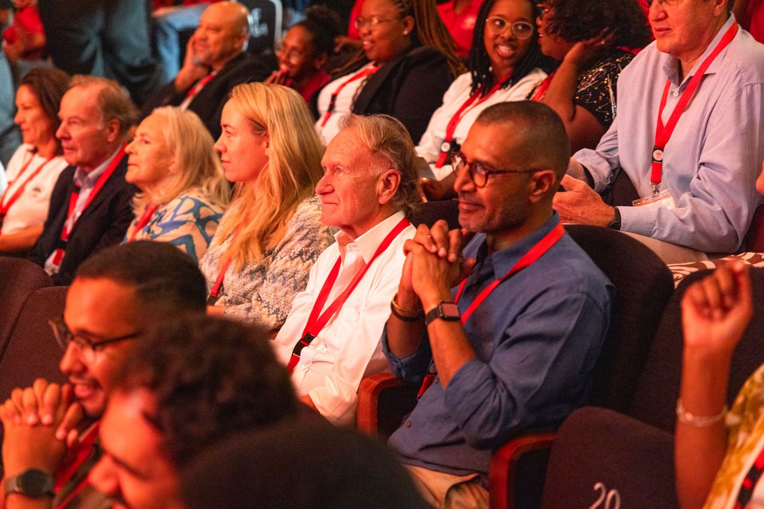 Diverse crowd of spectators seated in theater-style seating under dramatic red lighting.