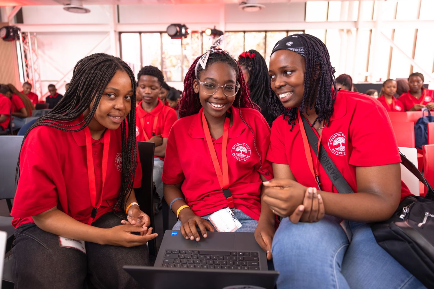 Three students in matching red shirts share a laugh while working on a laptop during an event.