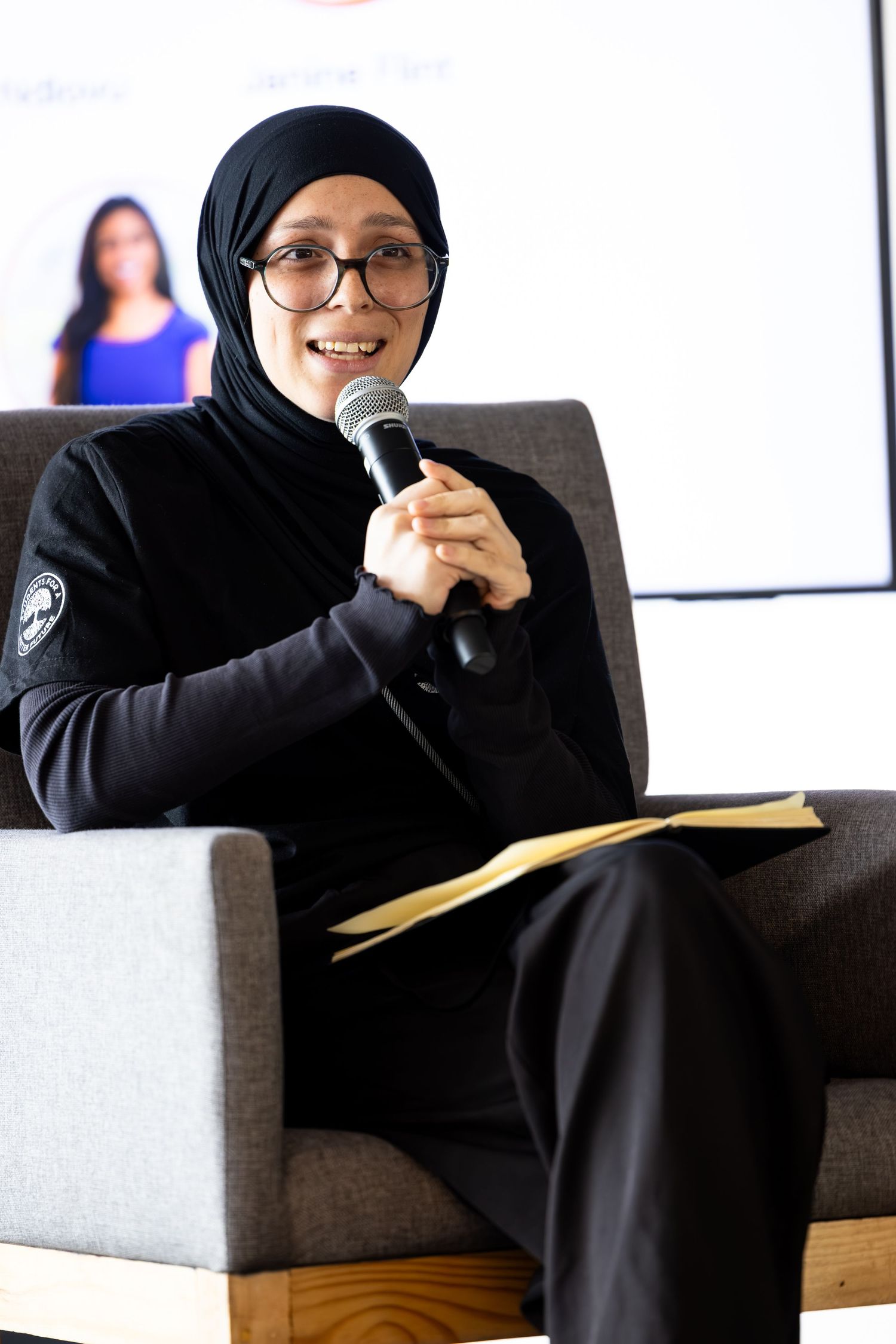 Person in black attire sits comfortably on a gray couch during a speaking engagement.