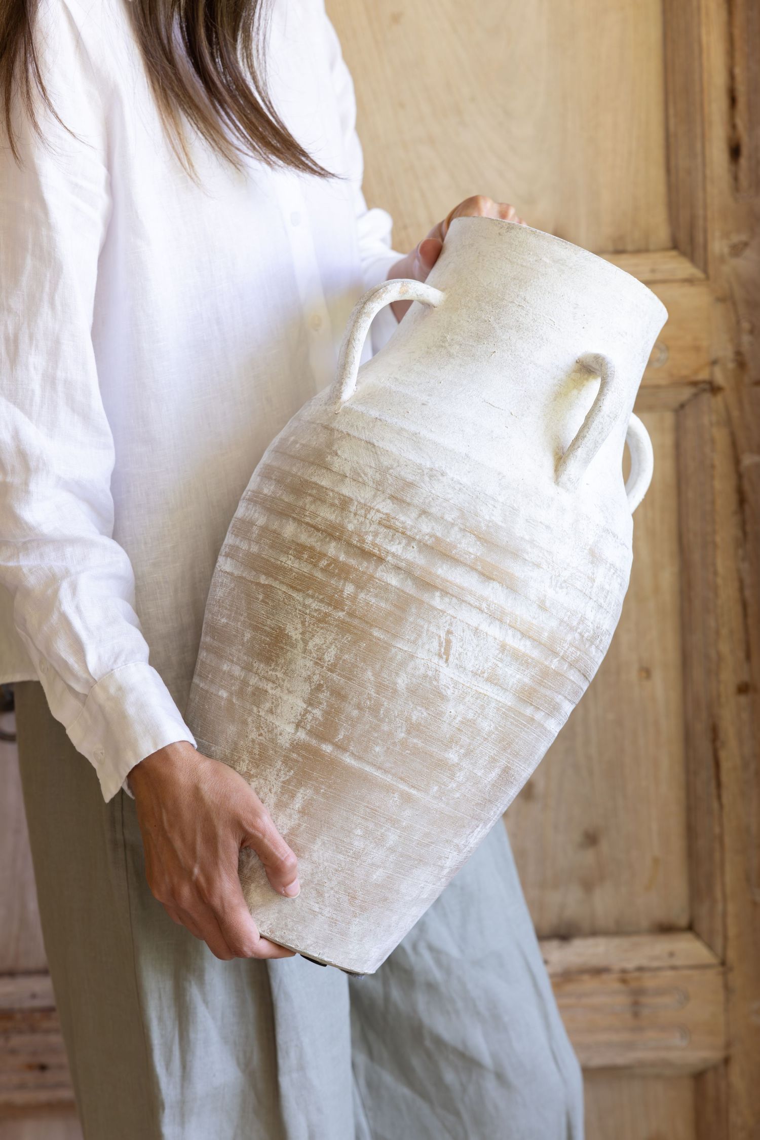 Person wearing white shirt holding large weathered antique ceramic amphora vase against rustic wooden background.