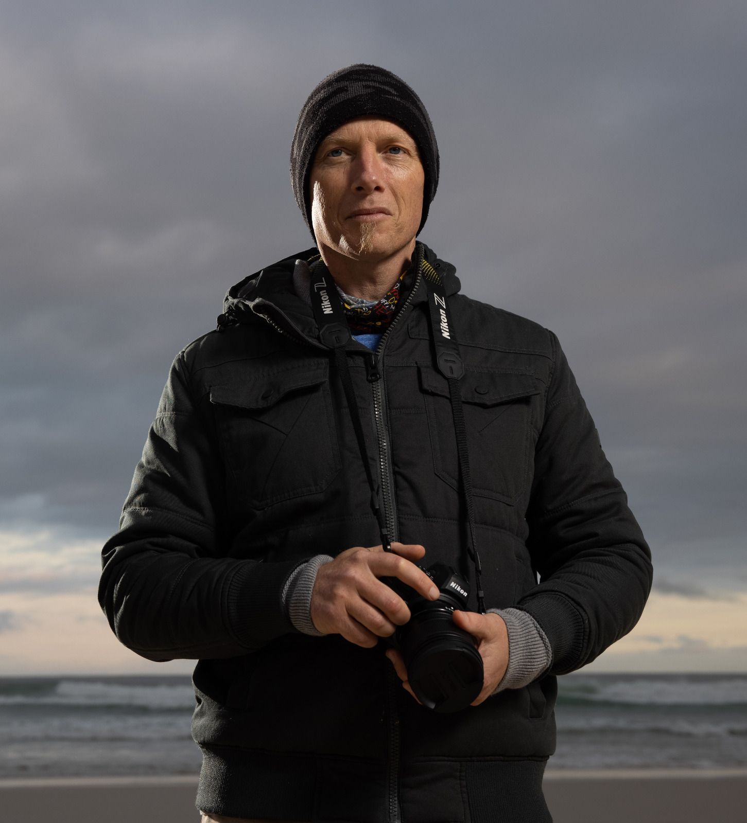 Person in black winter coat and beanie stands on beach holding camera against dramatic cloudy sky.
