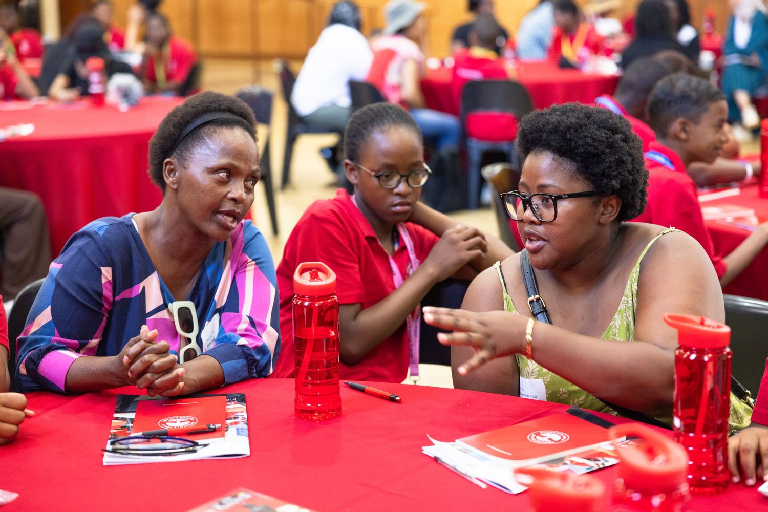 Participants in red shirts engage in group discussions at tables with learning materials.