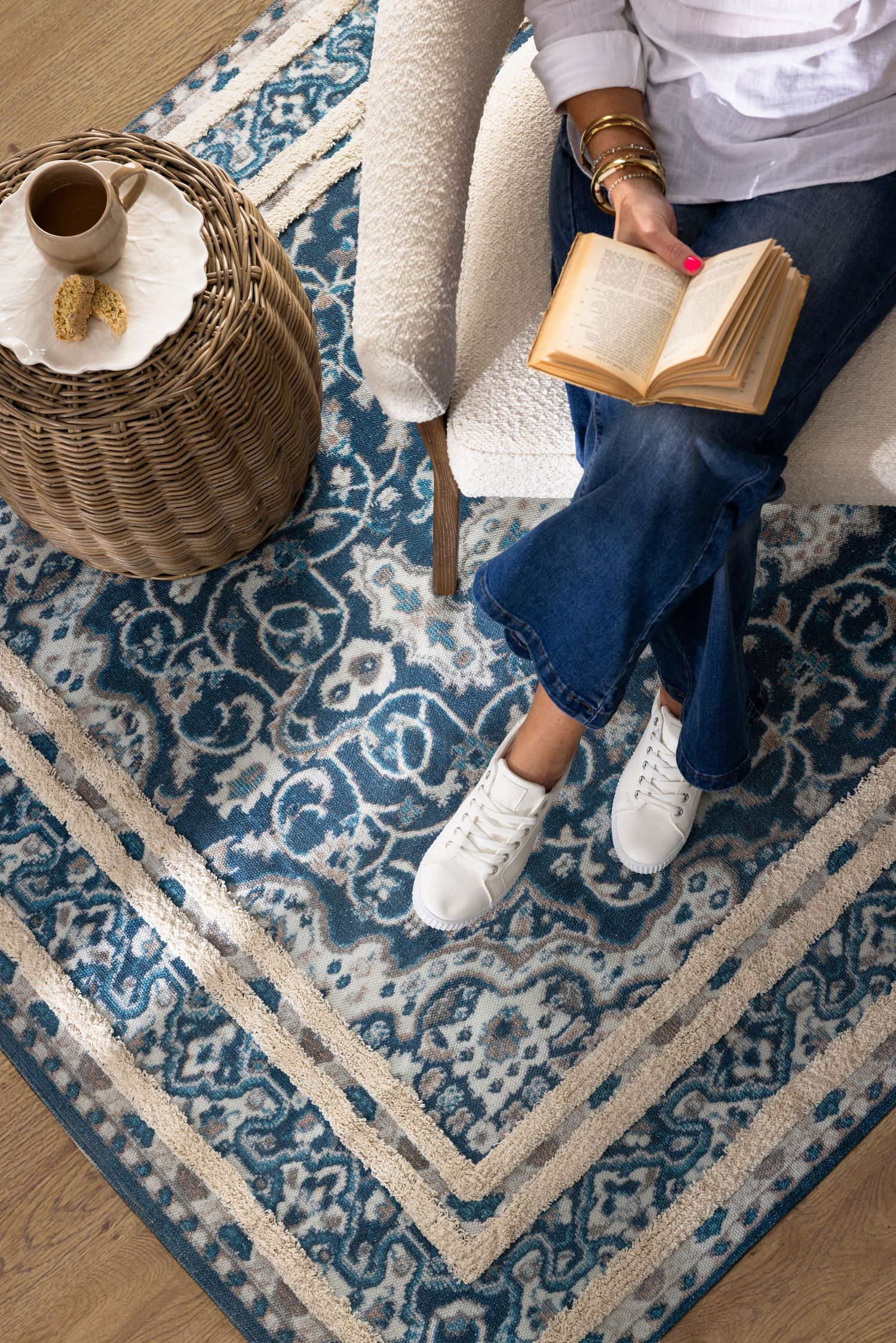 Reading nook scene with blue patterned rug, woven lamp, and open book against hardwood flooring.