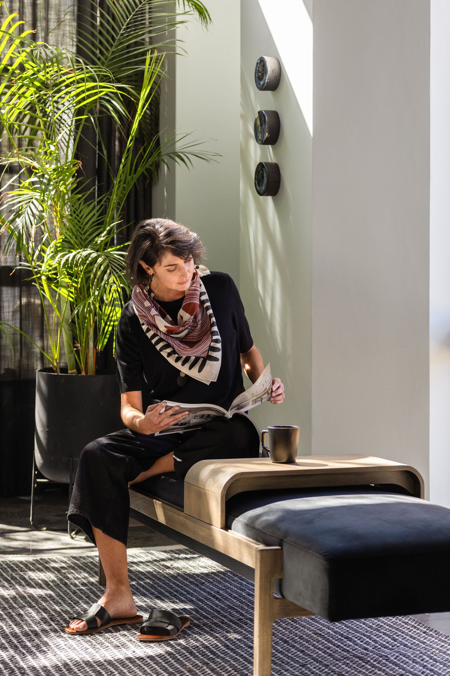 Person relaxes with reading material in a modern sunlit room with tropical plants and stylish bench seating.