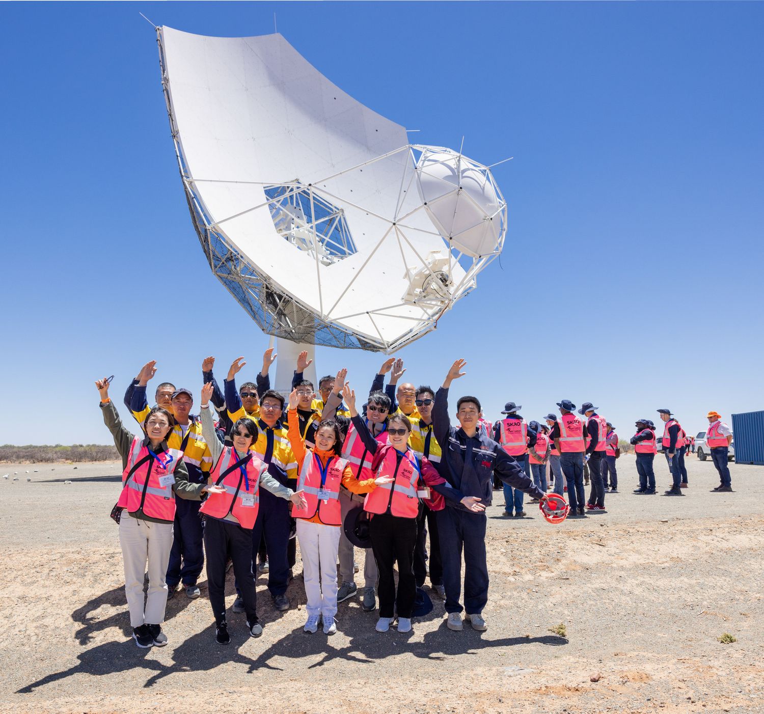 Large group in pink safety vests poses in front of white satellite dish equipment.