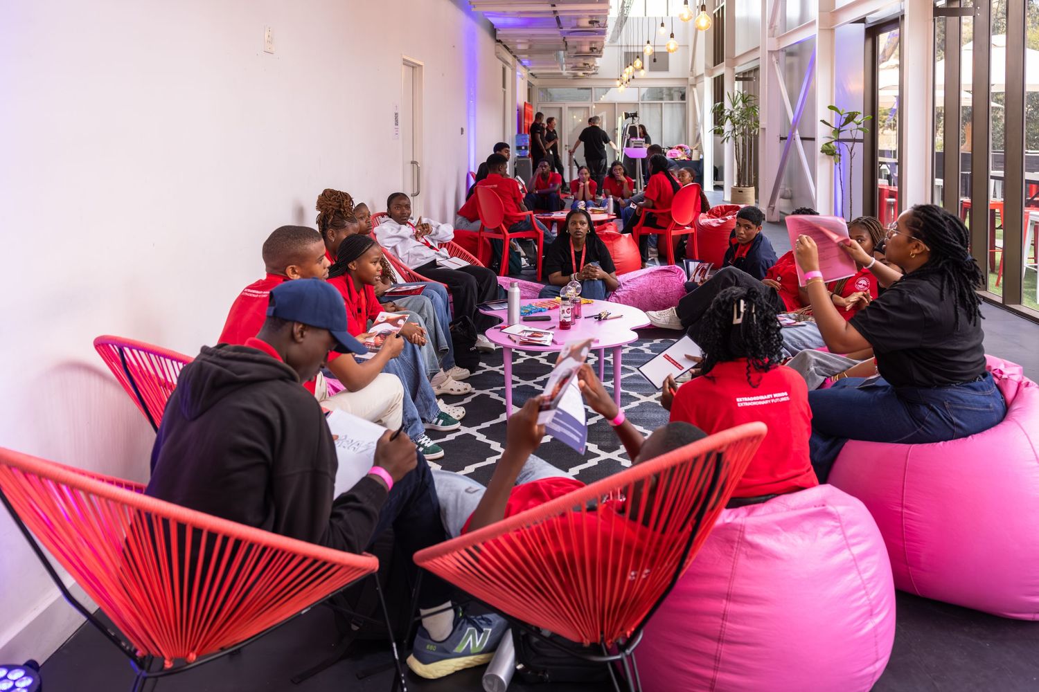 A row of modern red acapulco chairs with pink cushions line up against a white wall in an indoor space.