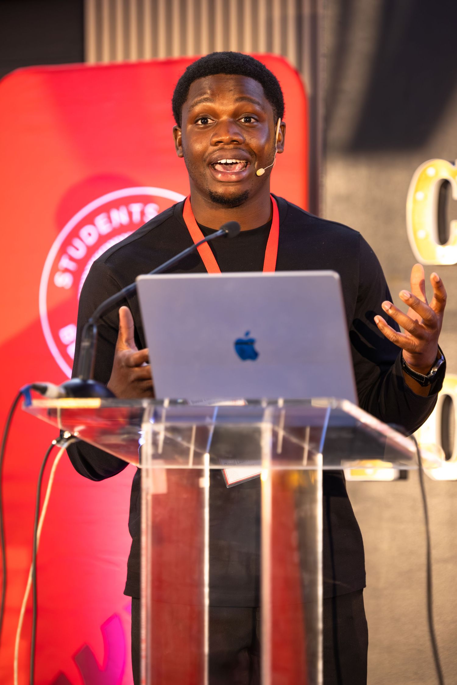 A speaker gestures while presenting behind clear podiums with a red-lit backdrop.