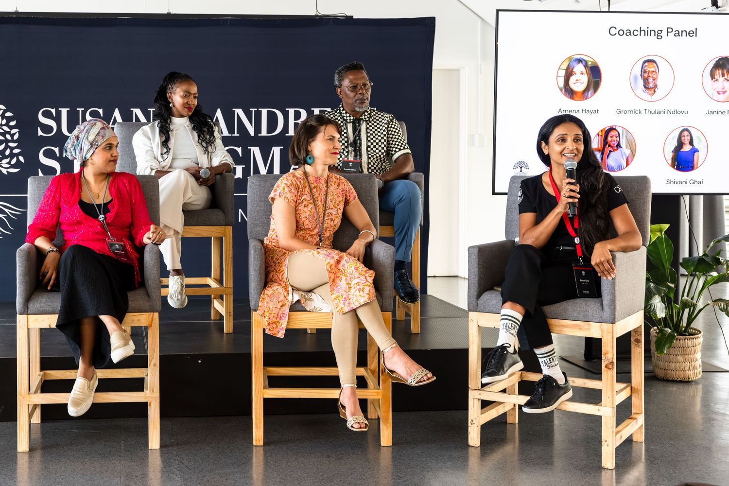 Row of people seated on wooden chairs against a branded backdrop at a professional event.