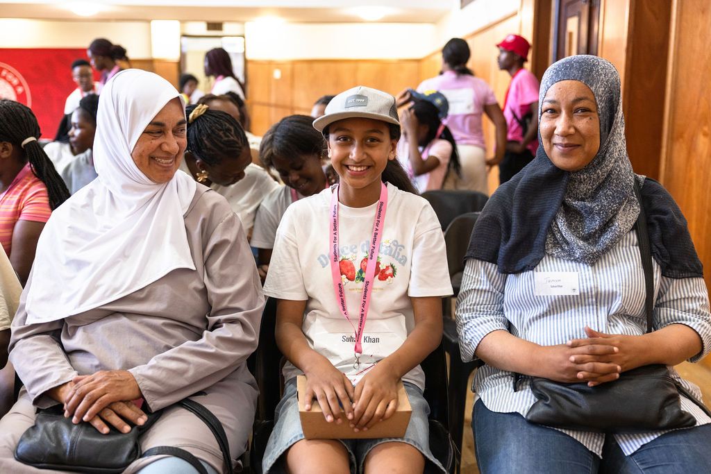 A group of people in hijabs and casual attire sit together smiling and conversing in a community gathering.