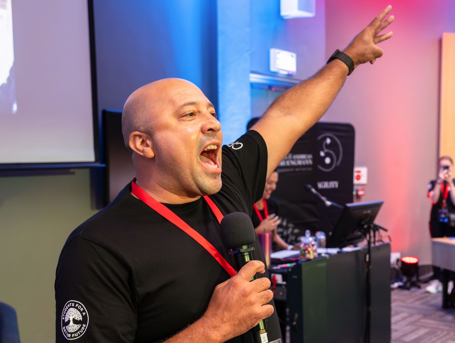 A speaker delivers an energetic presentation on stage at a business conference with blue and pink lighting.