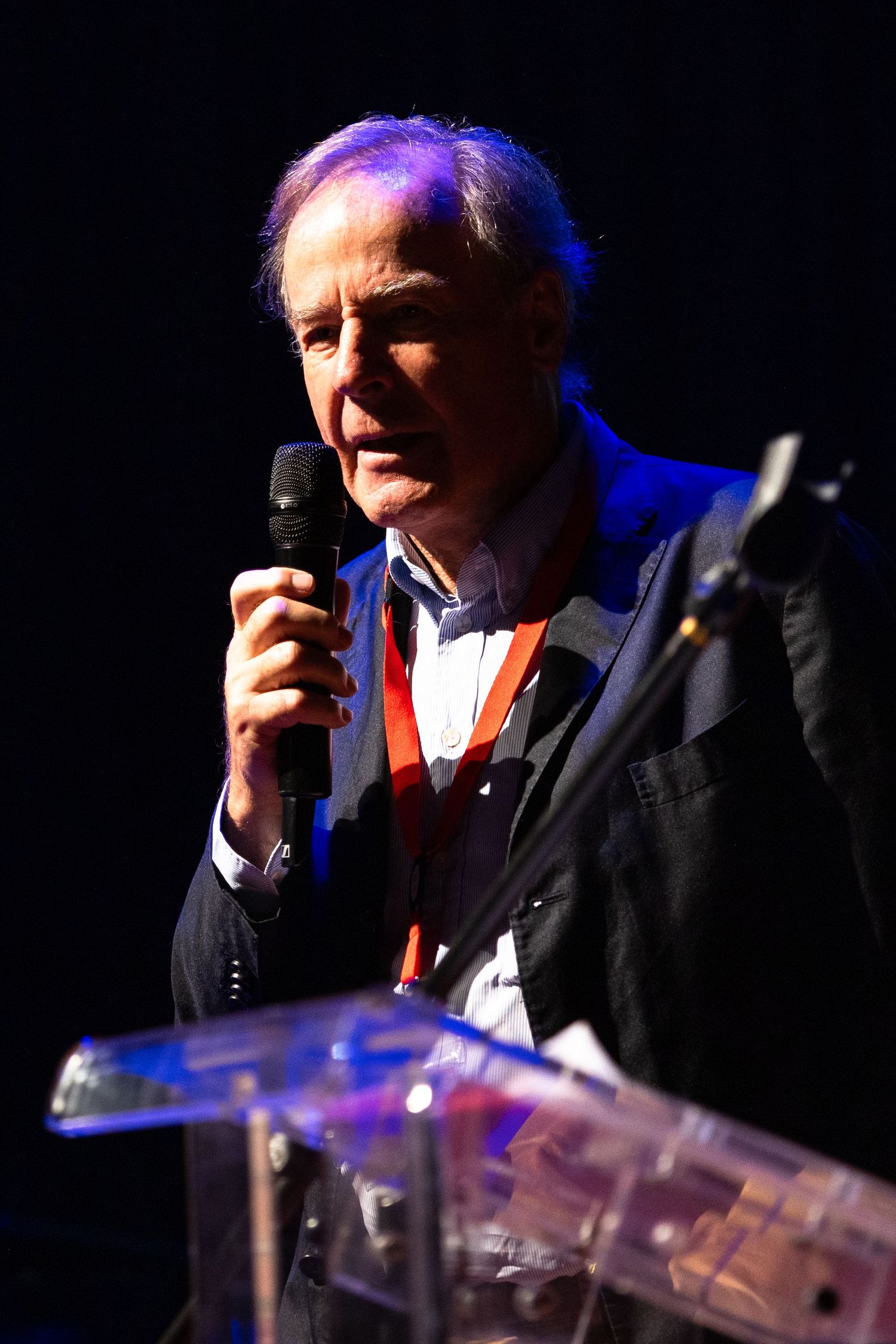 A speaker in a dark suit addresses an audience from behind a clear podium with dramatic stage lighting.