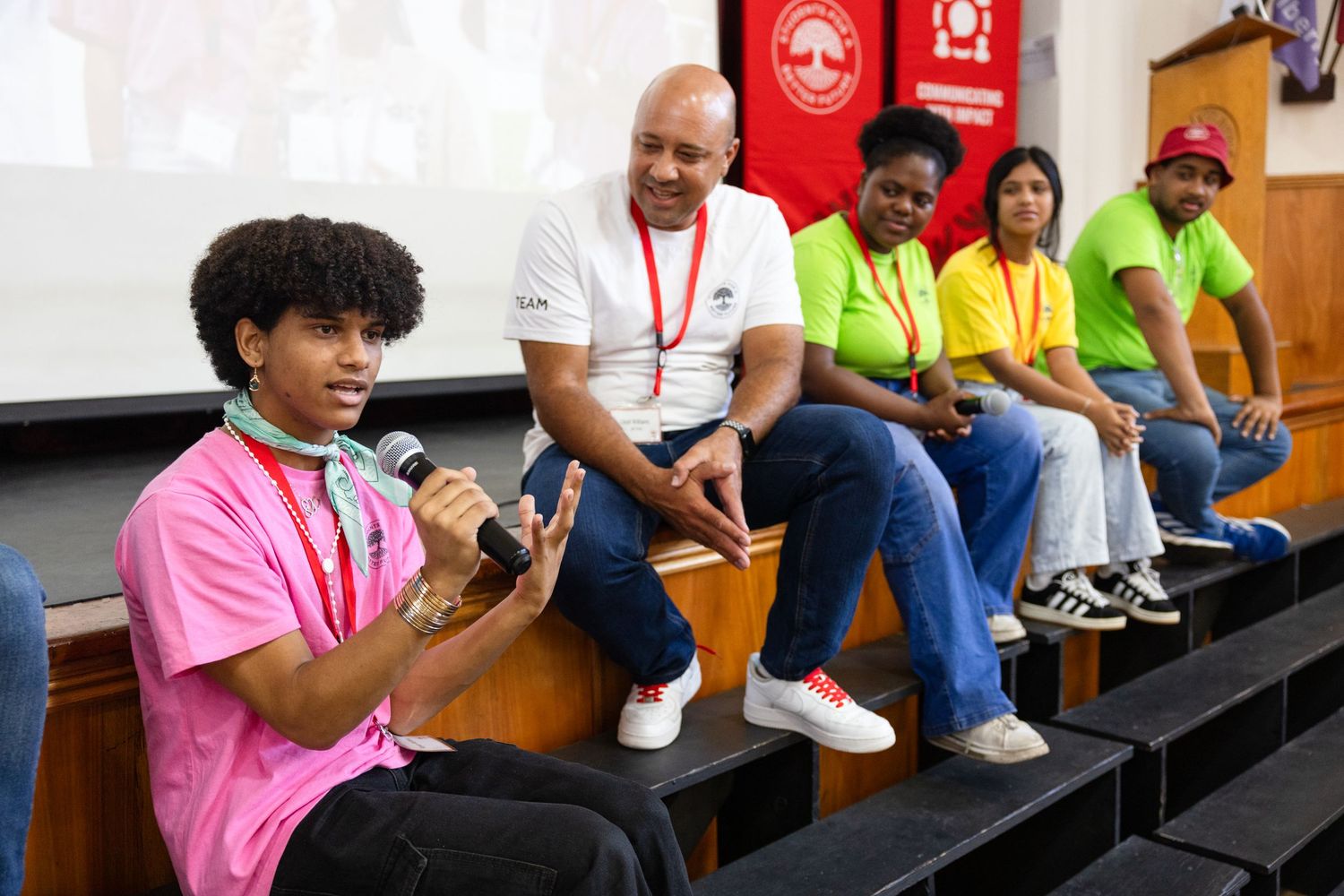 Group sits on bleacher steps during an indoor event with red banners visible.