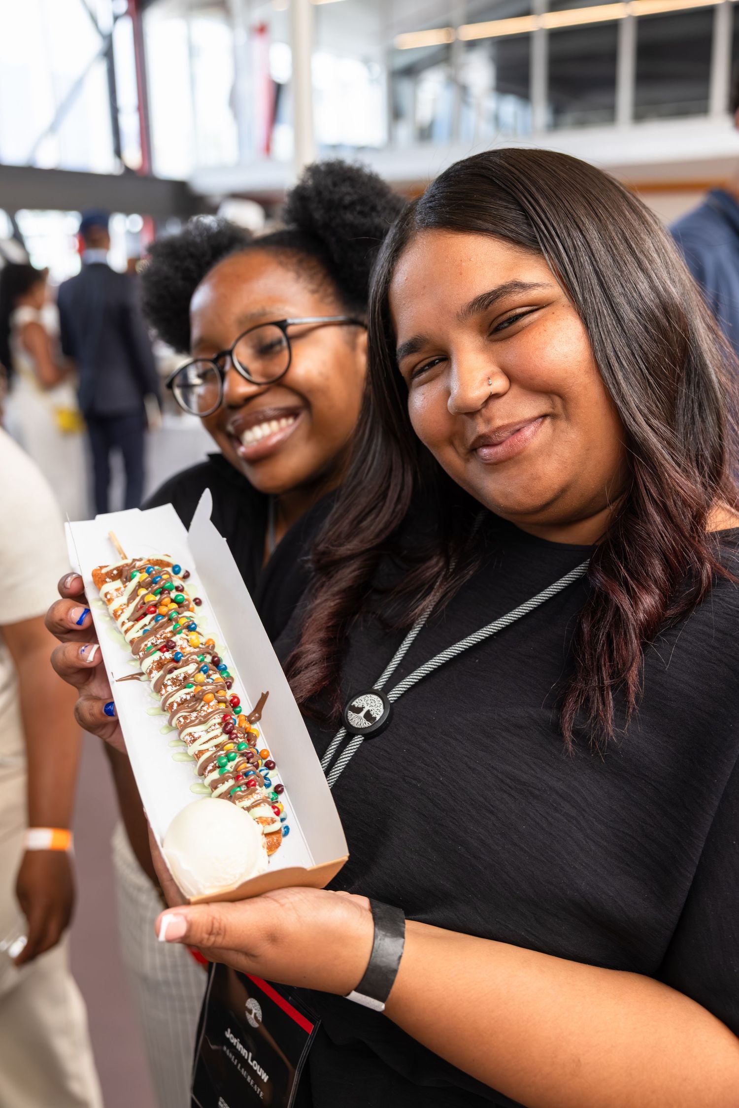 Two friends smile while holding a decorated hot dog with colorful toppings at a food event.