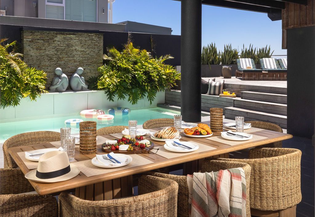 Outdoor dining area with wicker chairs and wooden table set against modern architectural backdrop and lush greenery.