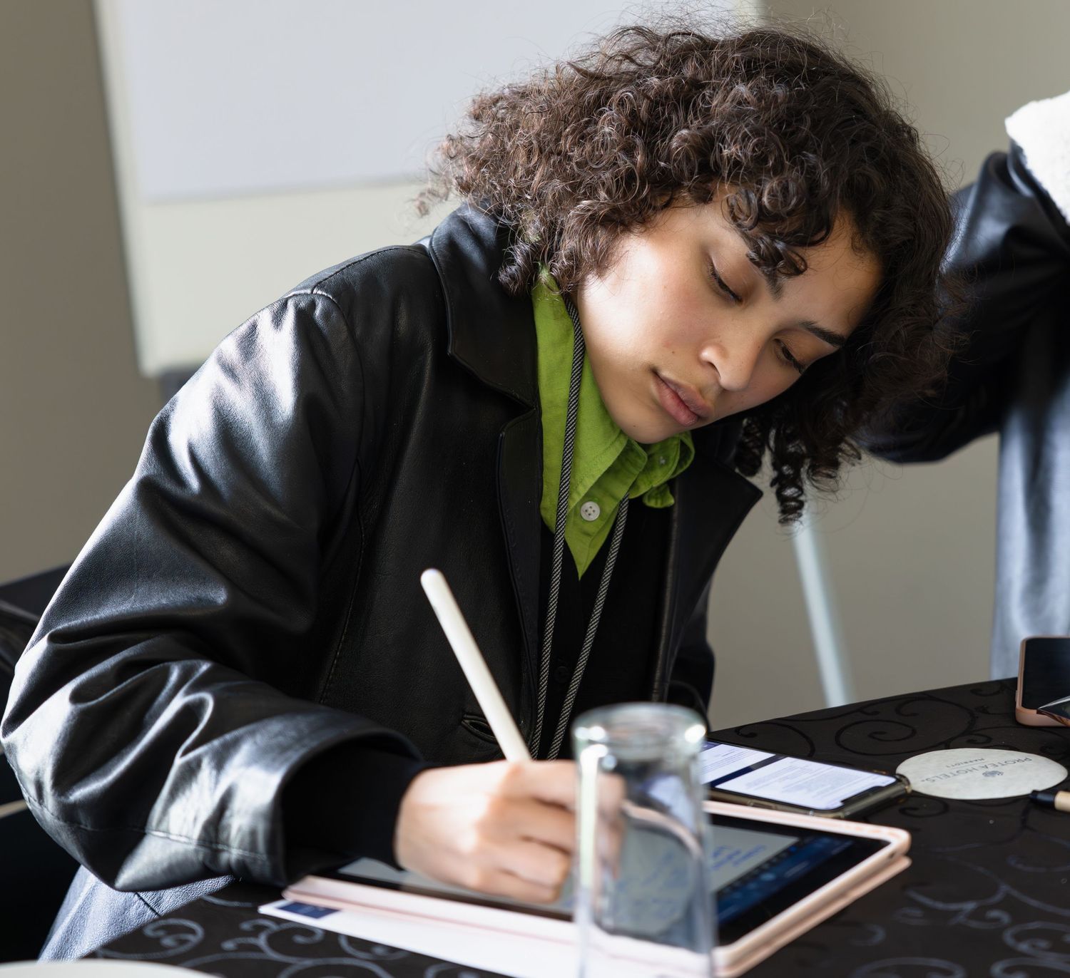 Student in black jacket focused on writing notes with a stylus on a digital tablet.