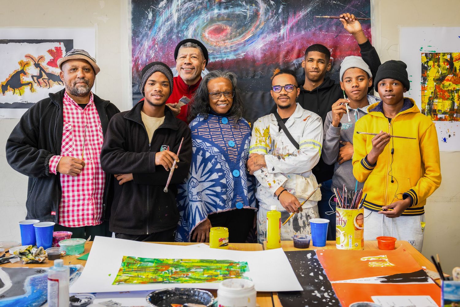 Artists gathered around artwork in a studio with a colorful galaxy painting in the background.