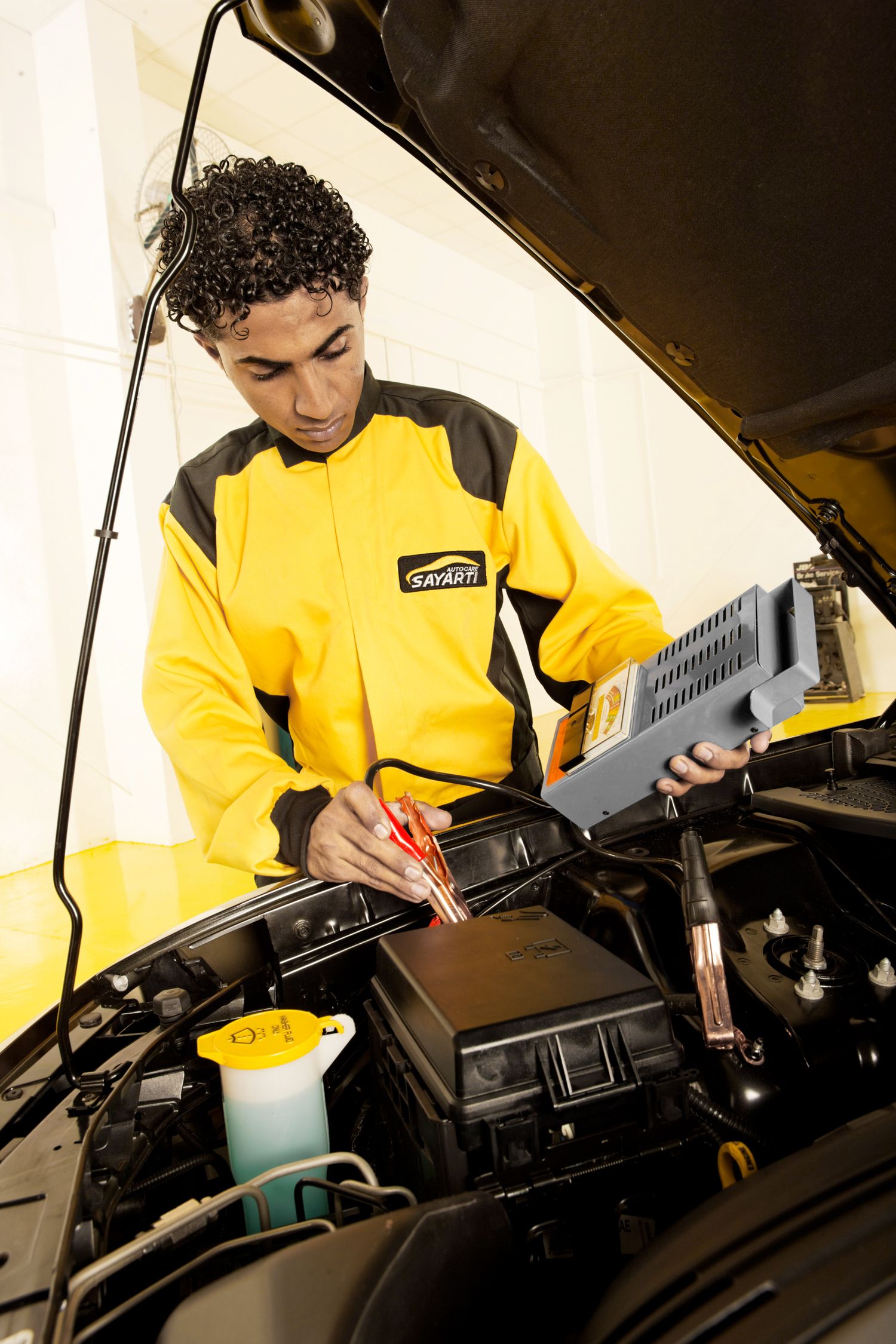 Automotive technician performing maintenance checks under vehicle hood while wearing yellow uniform.