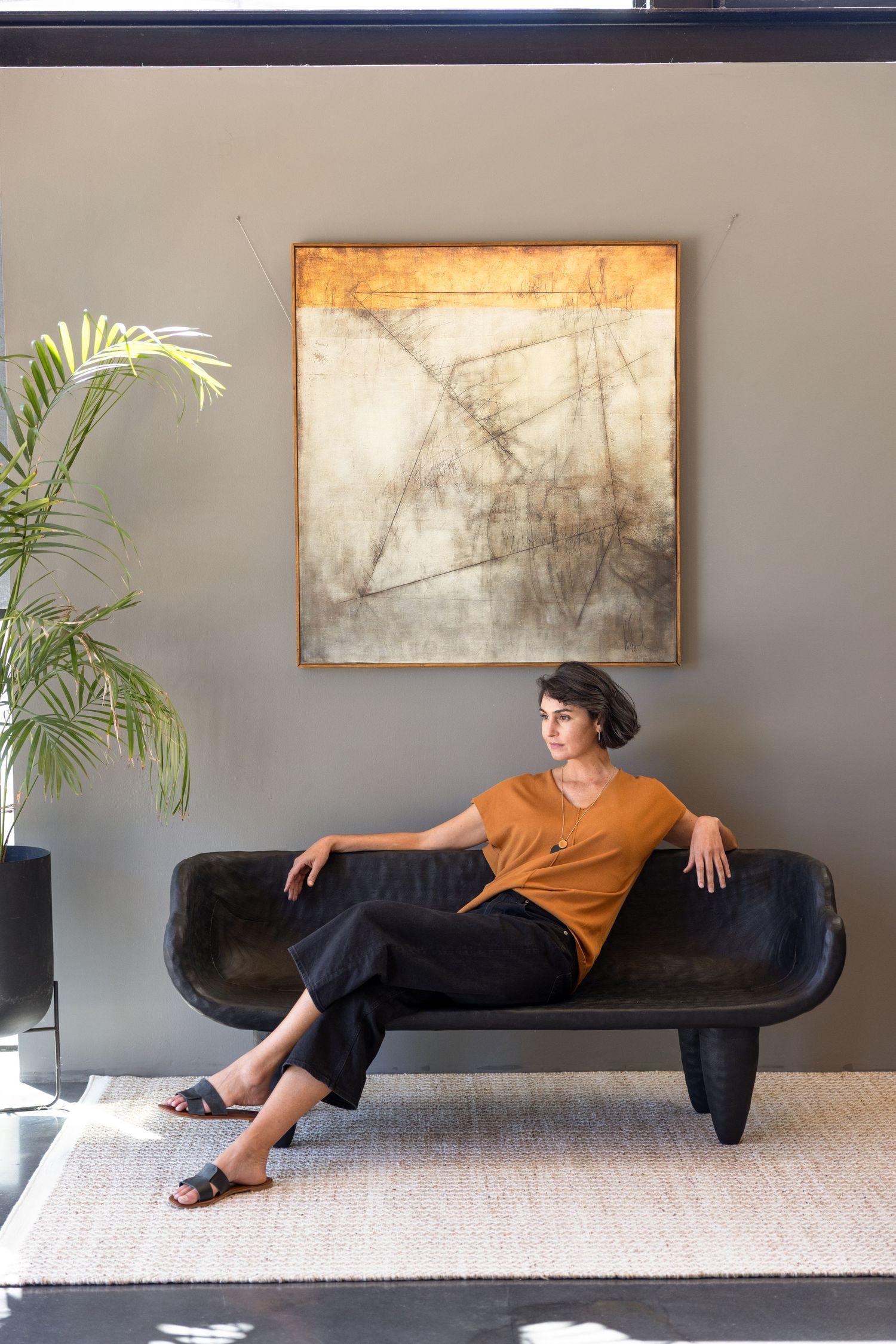 Interior design scene with person lounging on sleek black sofa beneath abstract artwork and palm plant.