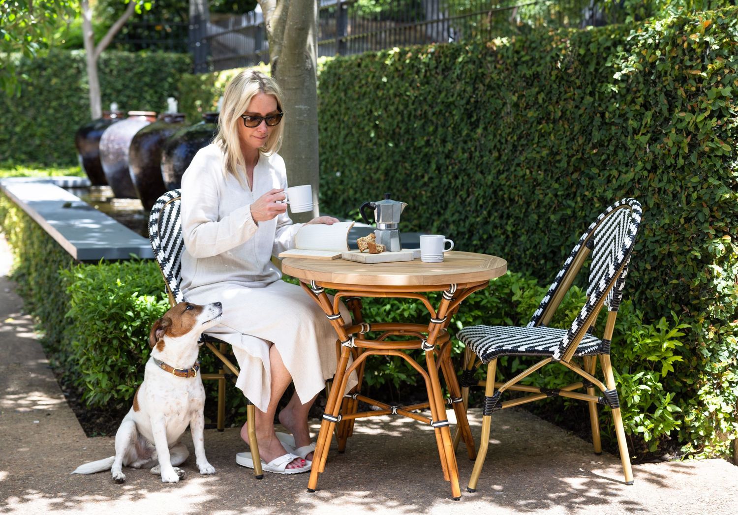 Someone sits at a charming outdoor cafe table with a small white dog beside them in a lush garden setting.