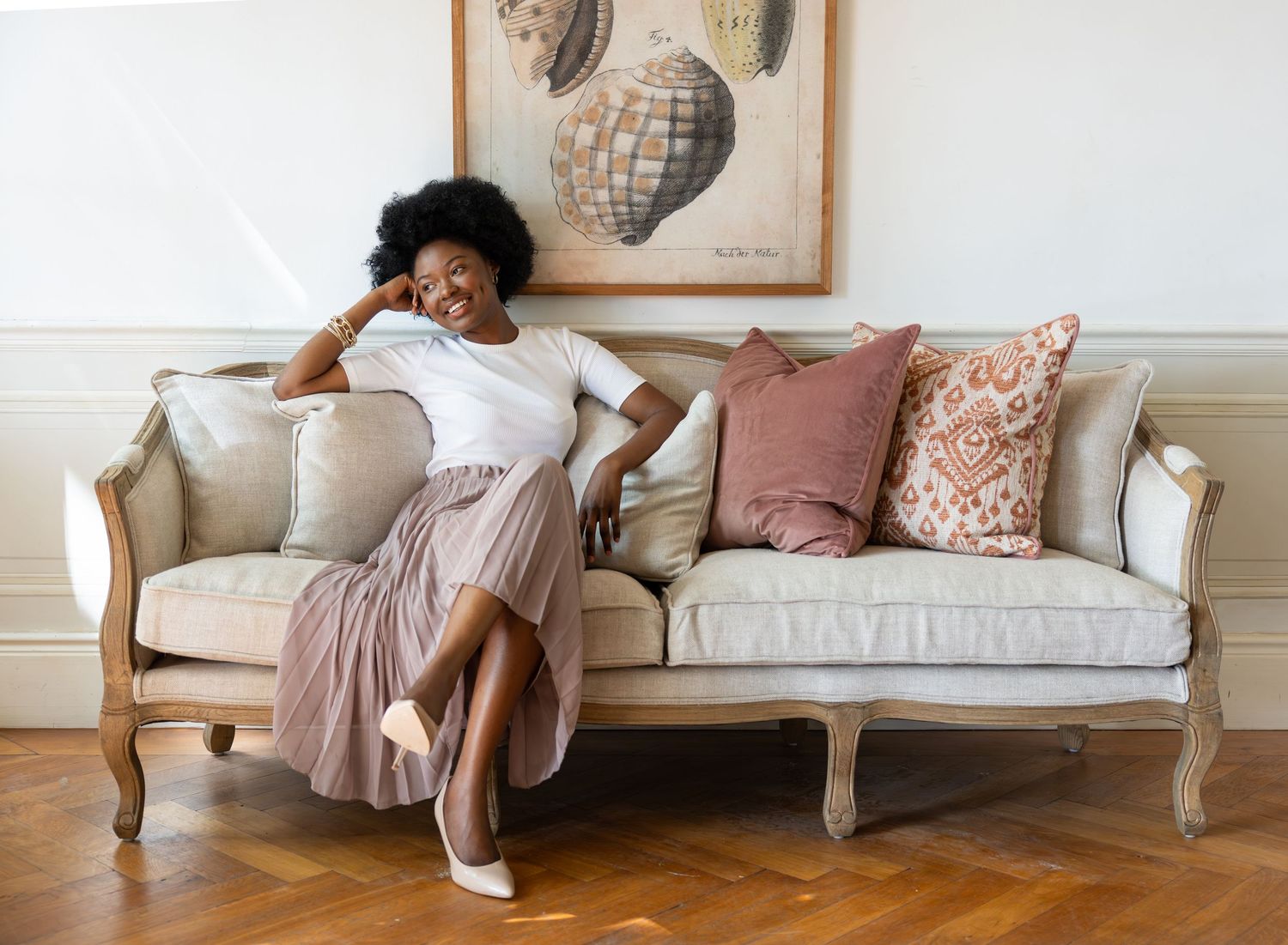 A person lounges casually on a vintage French-style sofa with pink and beige throw pillows beneath framed wall art.