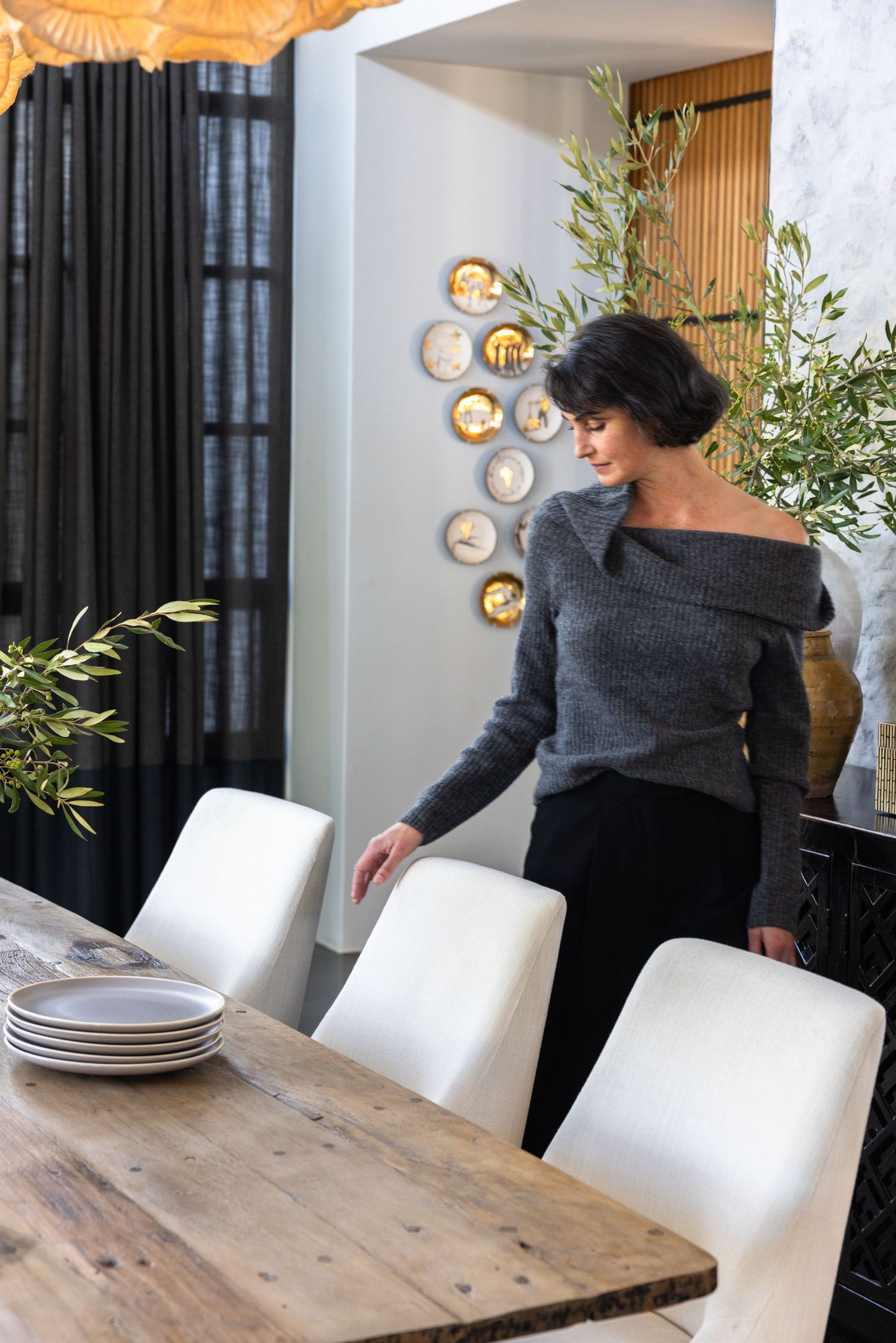A person arranging place settings on a rustic wooden dining table with white chairs and modern wall sconces.