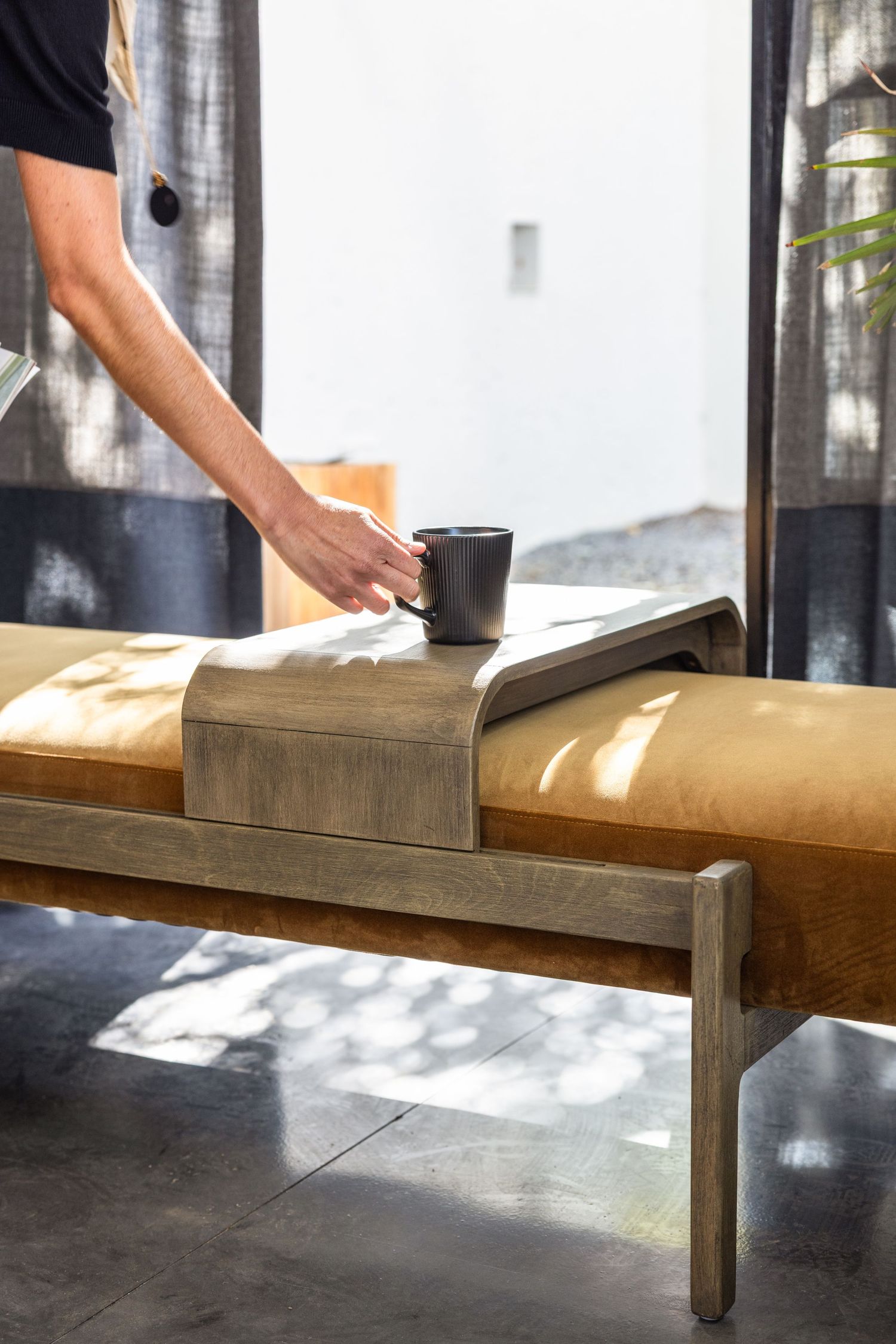 Hand placing black coffee cup on wooden bench tray with sunlight streaming through sheer curtains.