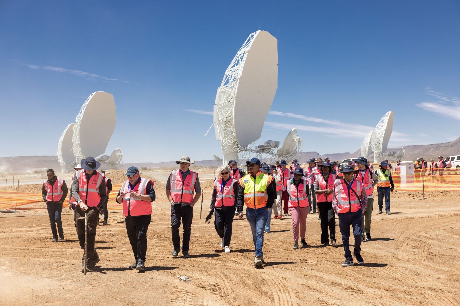 Group in safety vests walking past large white satellite dishes in desert setting.