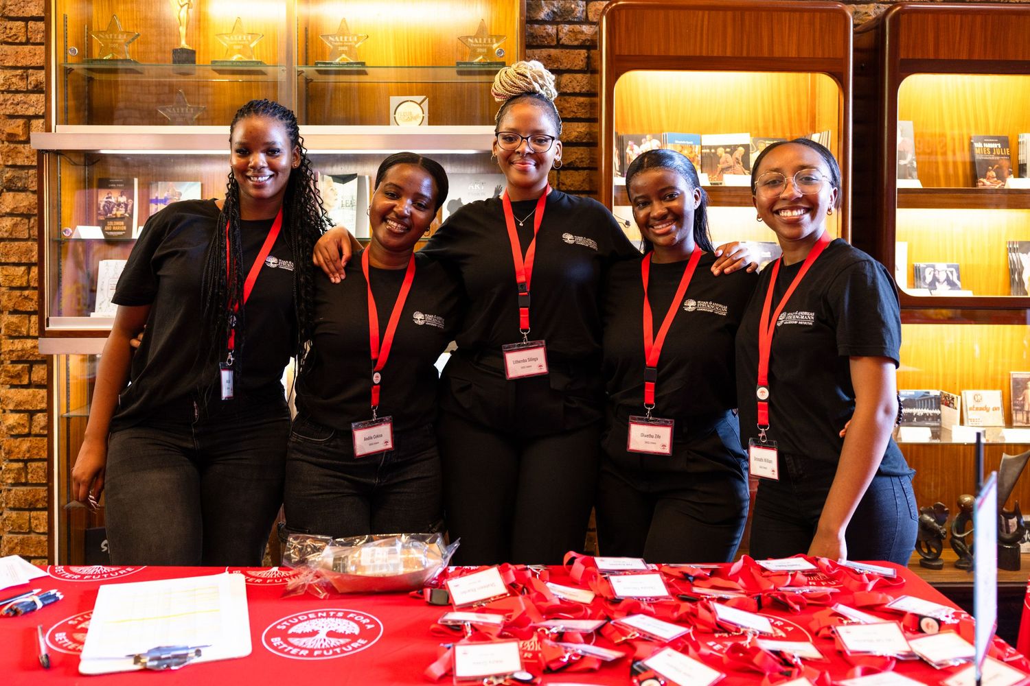 Staff members in black uniforms stand behind a red-clothed table at a special event.