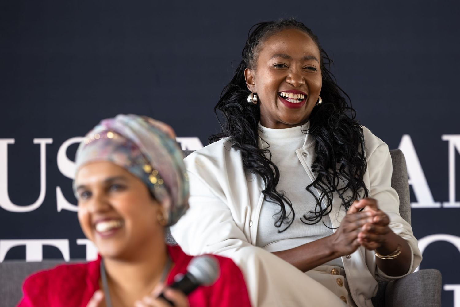 Two speakers share a lighthearted moment during a panel discussion against a dark background.