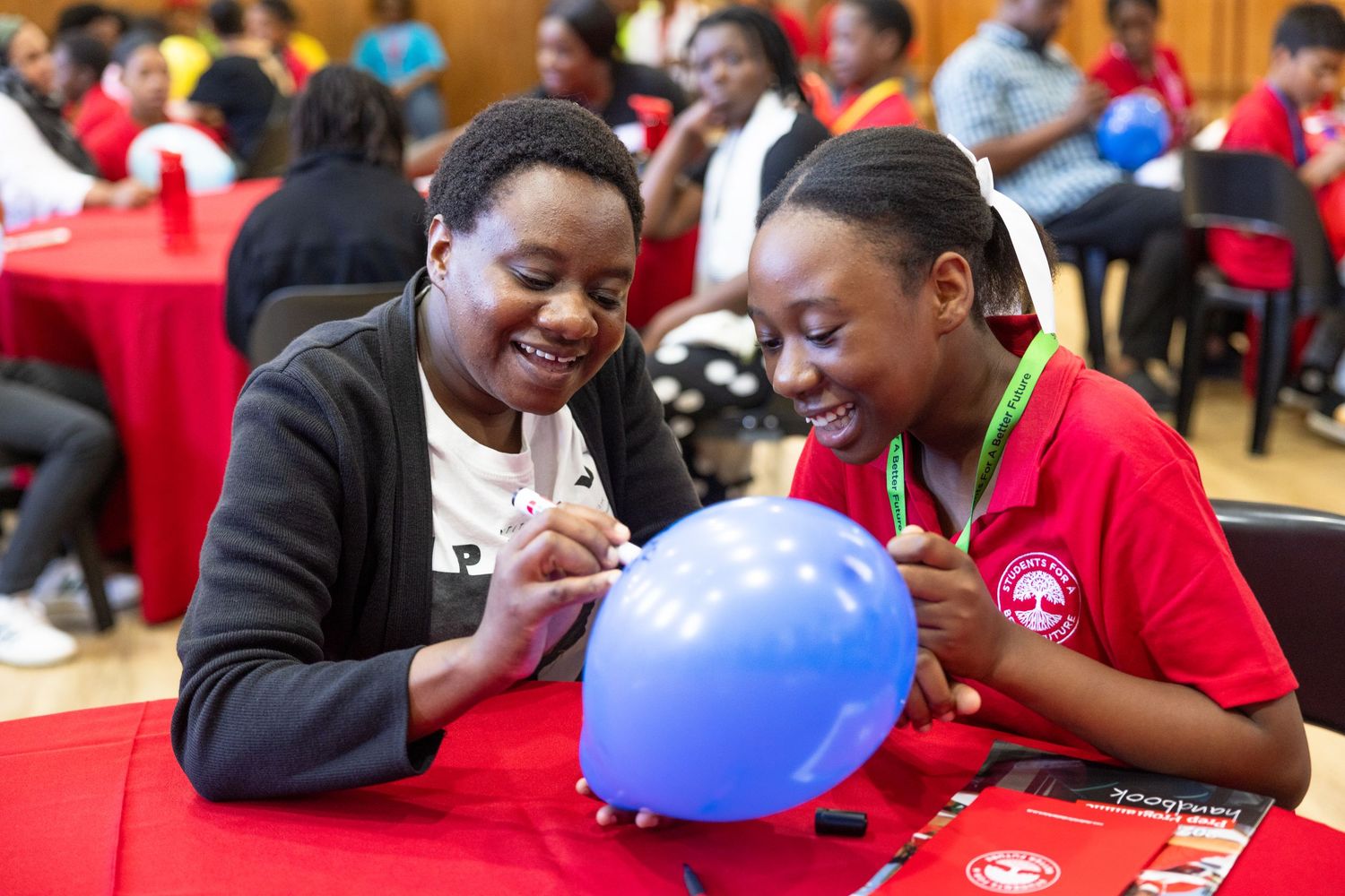 Students in red shirts smile and laugh while working together on a classroom activity with colorful balloons.