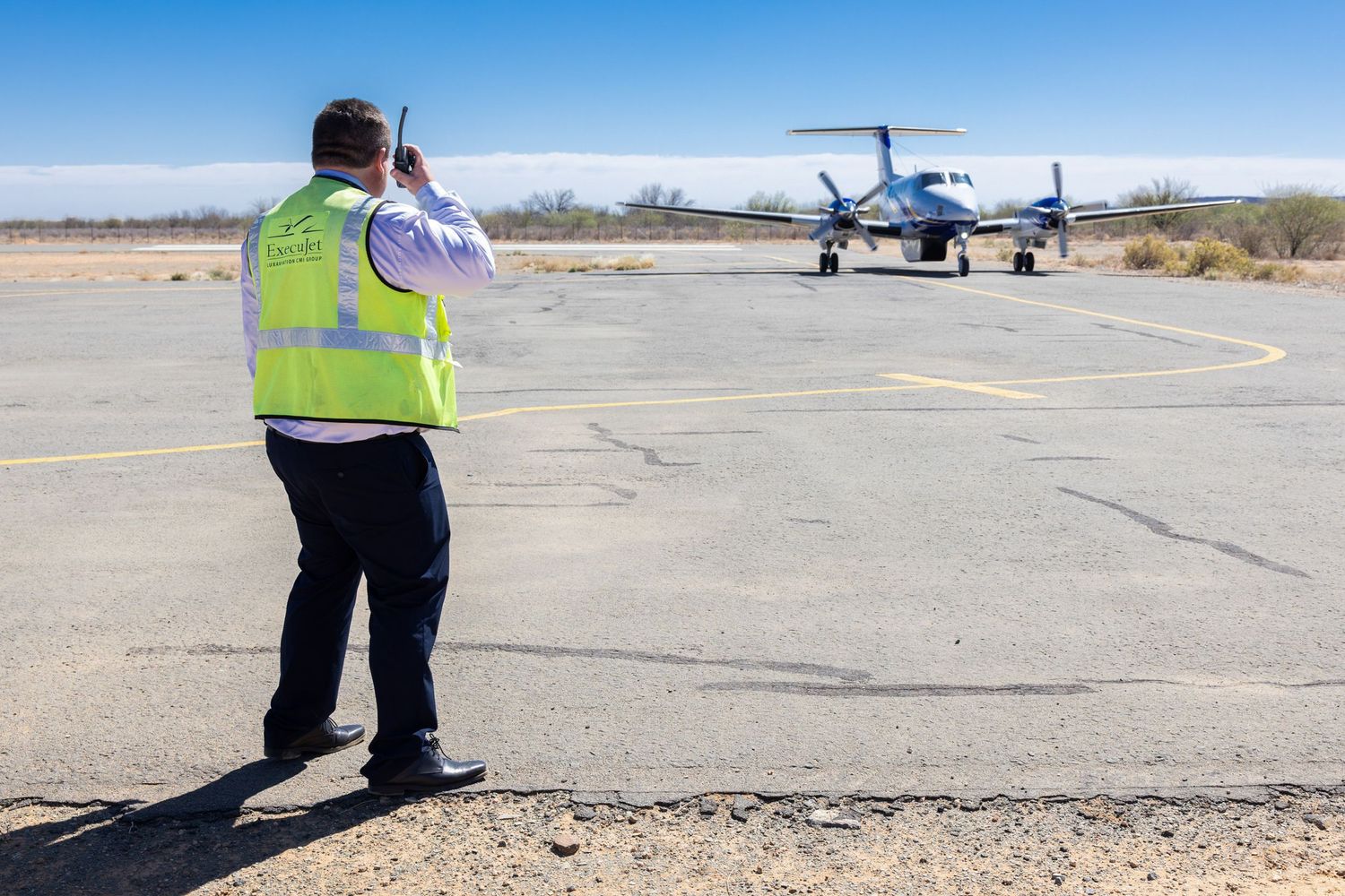 Airport ground crew member in safety vest photographs small aircraft on desert runway.