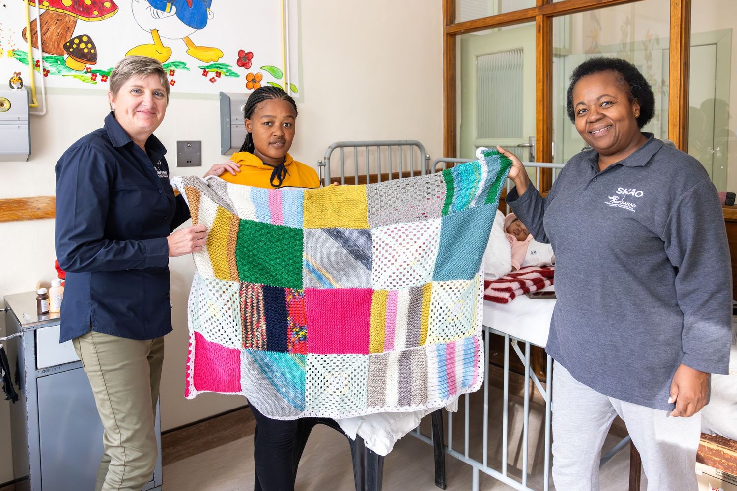 People holding up a handmade patchwork quilt with bright colored squares in a medical facility.