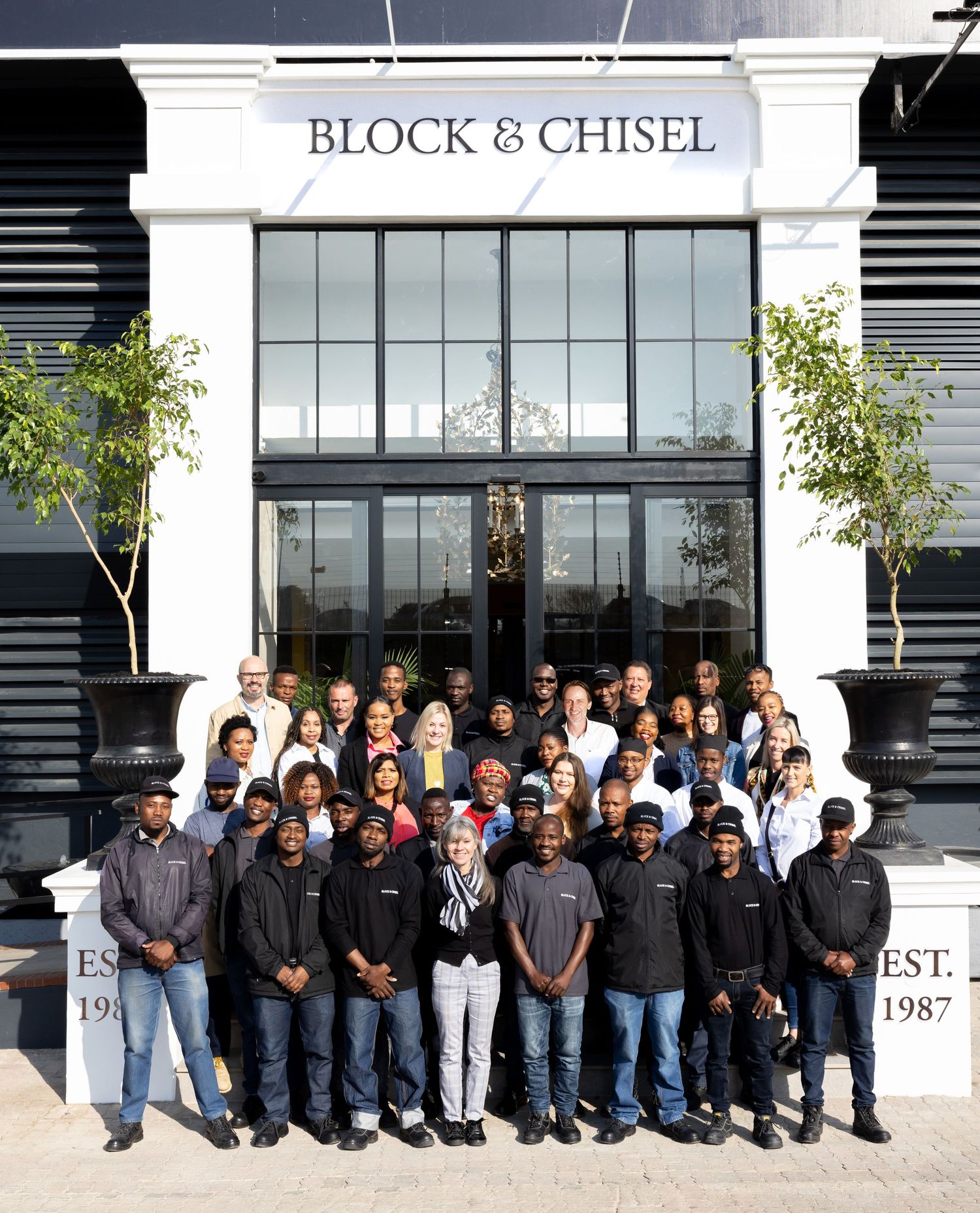 Large group poses in front of Block & Chisel storefront with black and white facade.