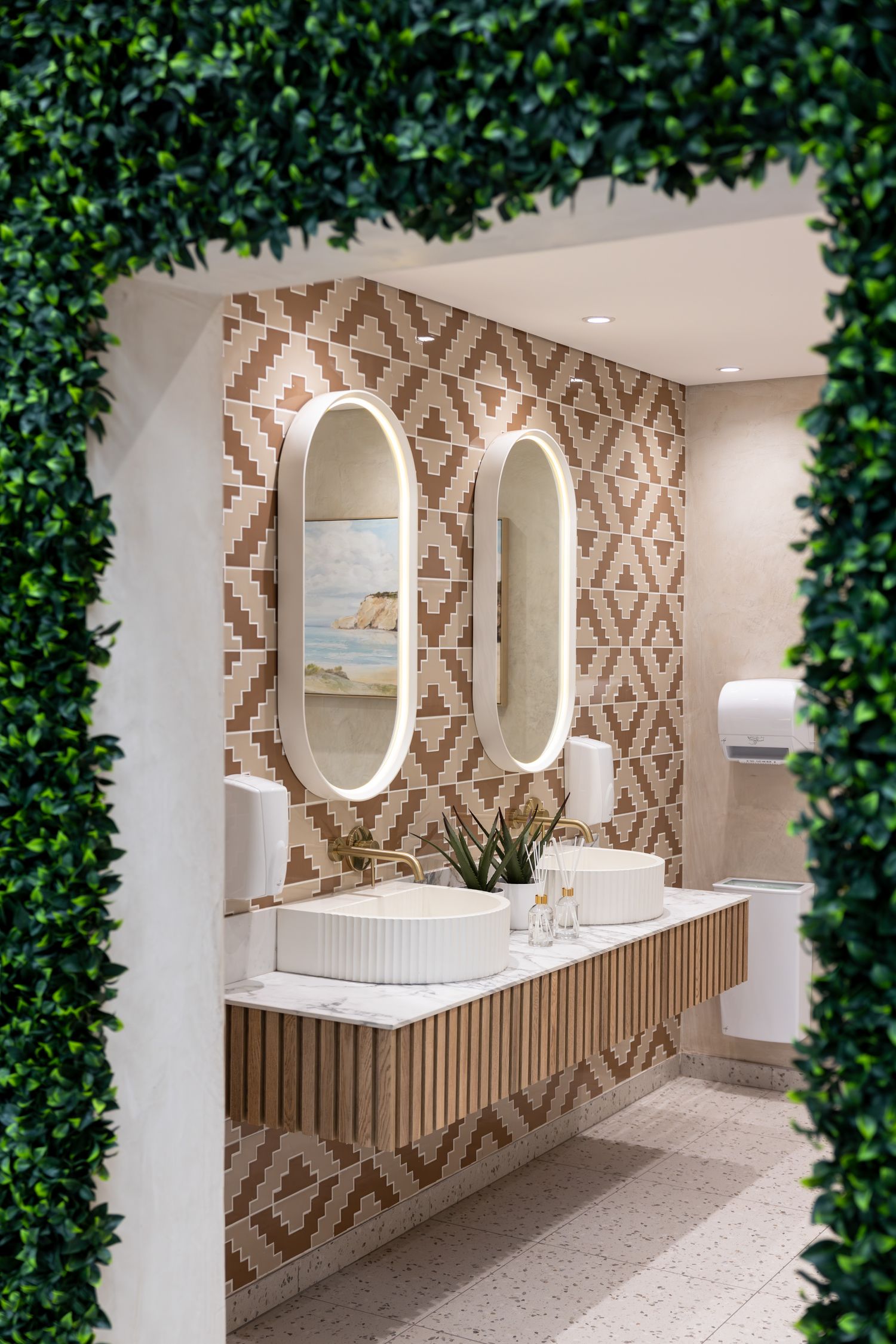 Luxurious bathroom display with oval mirrors and geometric wallpaper framed by decorative green foliage.